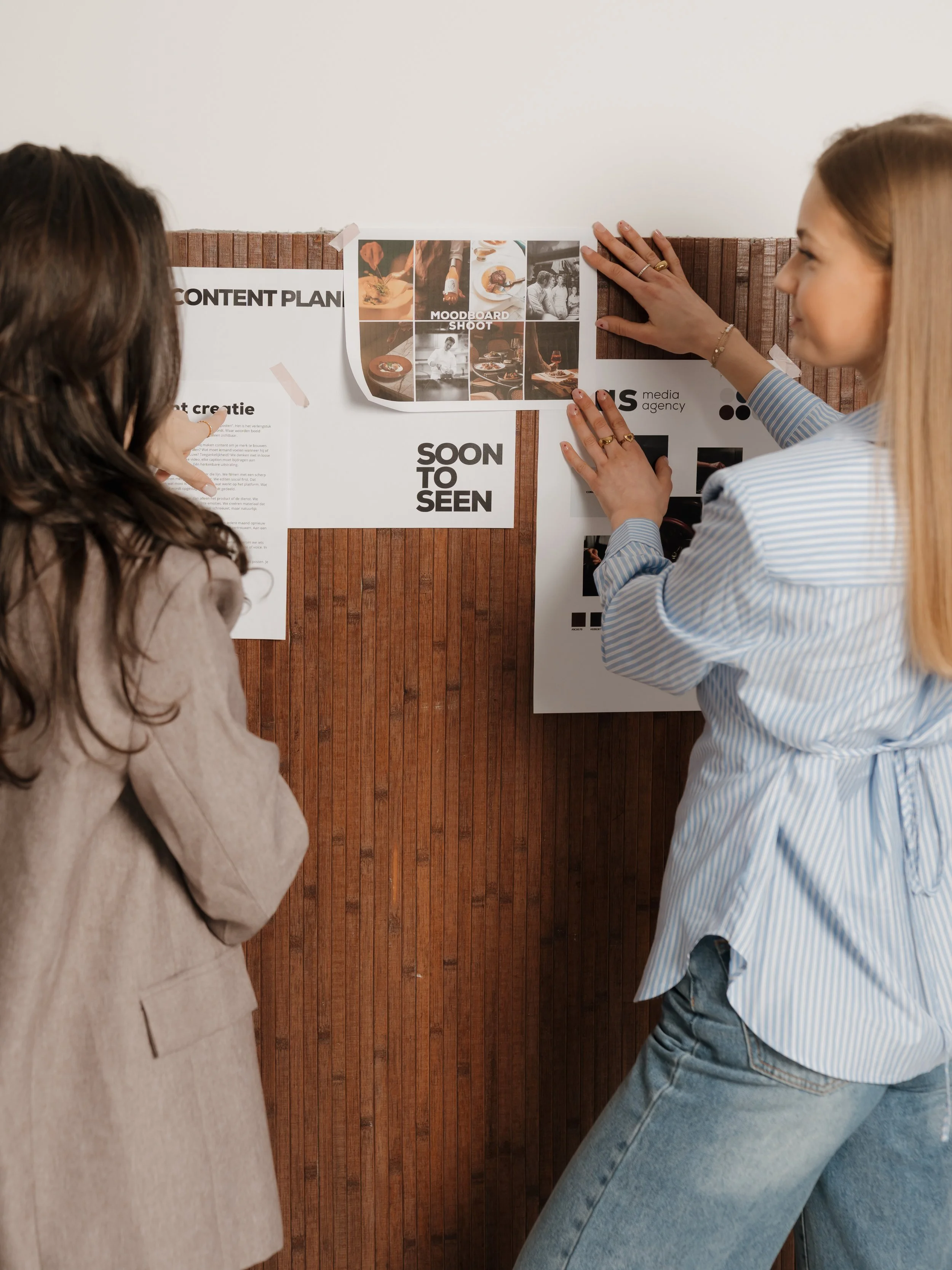 Two women looking at a bulletin board with papers and photographs tacked on it, in a discussion or planning setting.