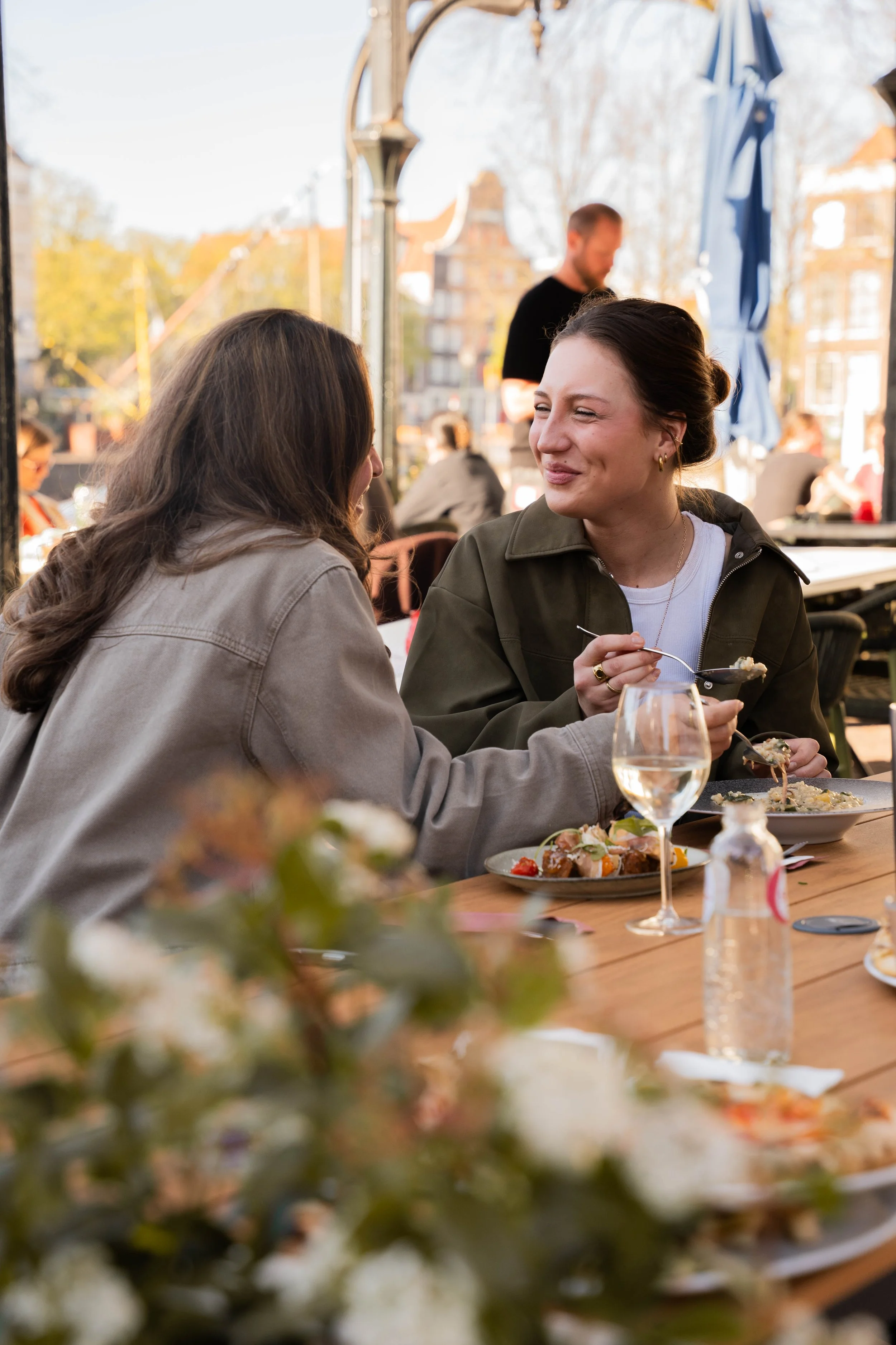 Two women are sitting at an outdoor restaurant table, smiling and enjoying a meal together. There are plates of food and a glass of white wine on the table. In the background, there are other people dining and a man standing near a blue umbrella.