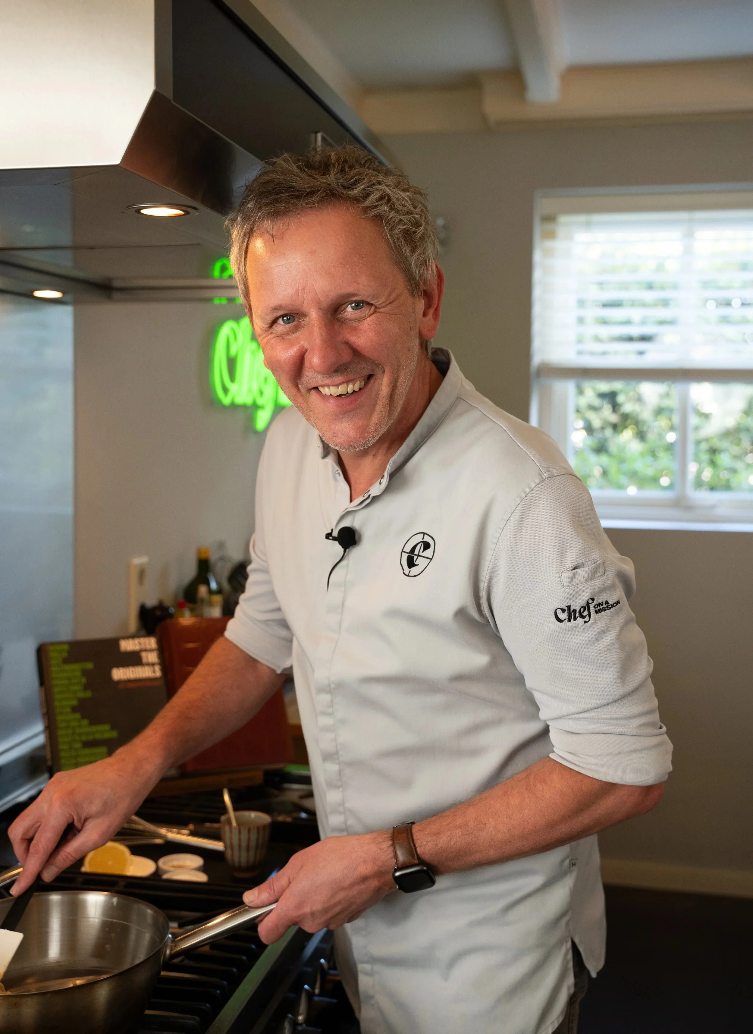 A smiling man in a white chef's coat cooking in a kitchen, with a neon green sign and a window in the background.