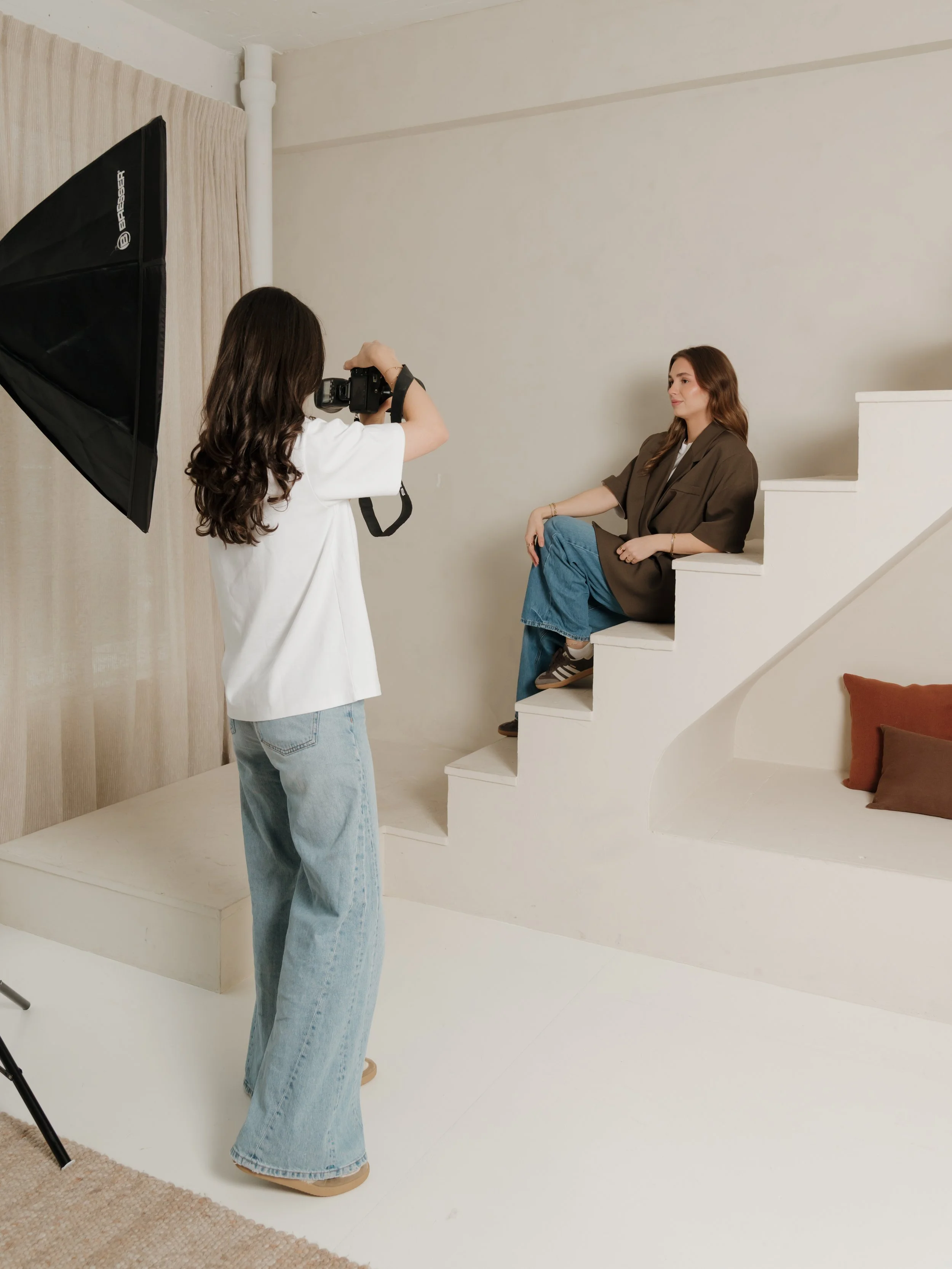 Woman with long brown hair sitting on white stairs during a photo shoot with a photographer taking her picture inside a studio.