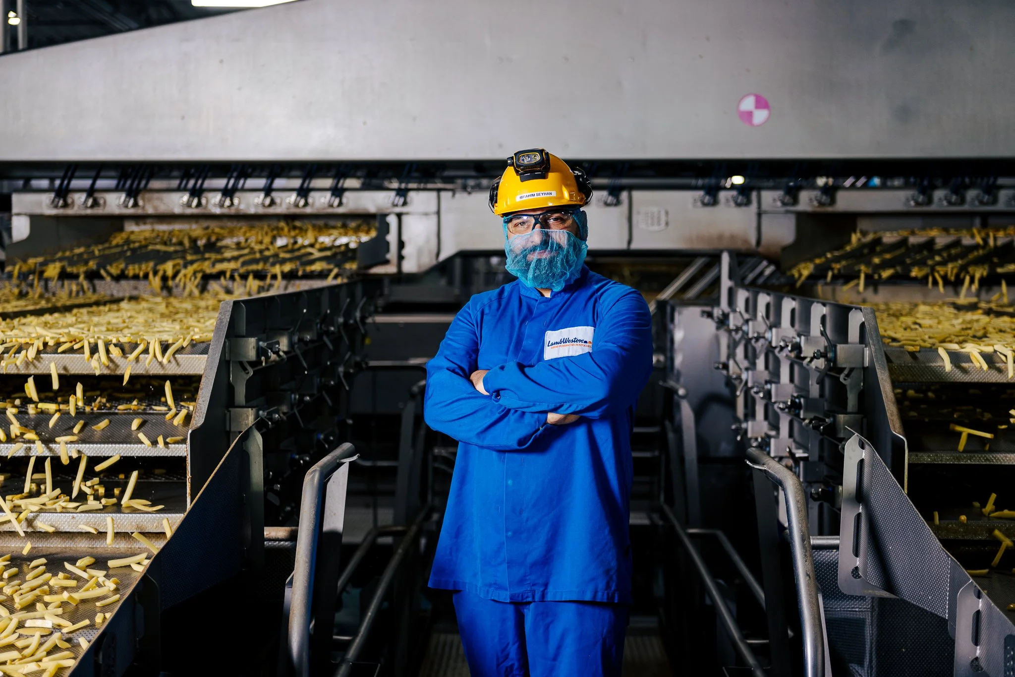 A man wearing a yellow safety helmet, blue work uniform, glasses, and a blue beard net stands with crossed arms in a food processing facility. In the background, conveyor belts are filled with sliced potatoes being processed for cooking or packaging.