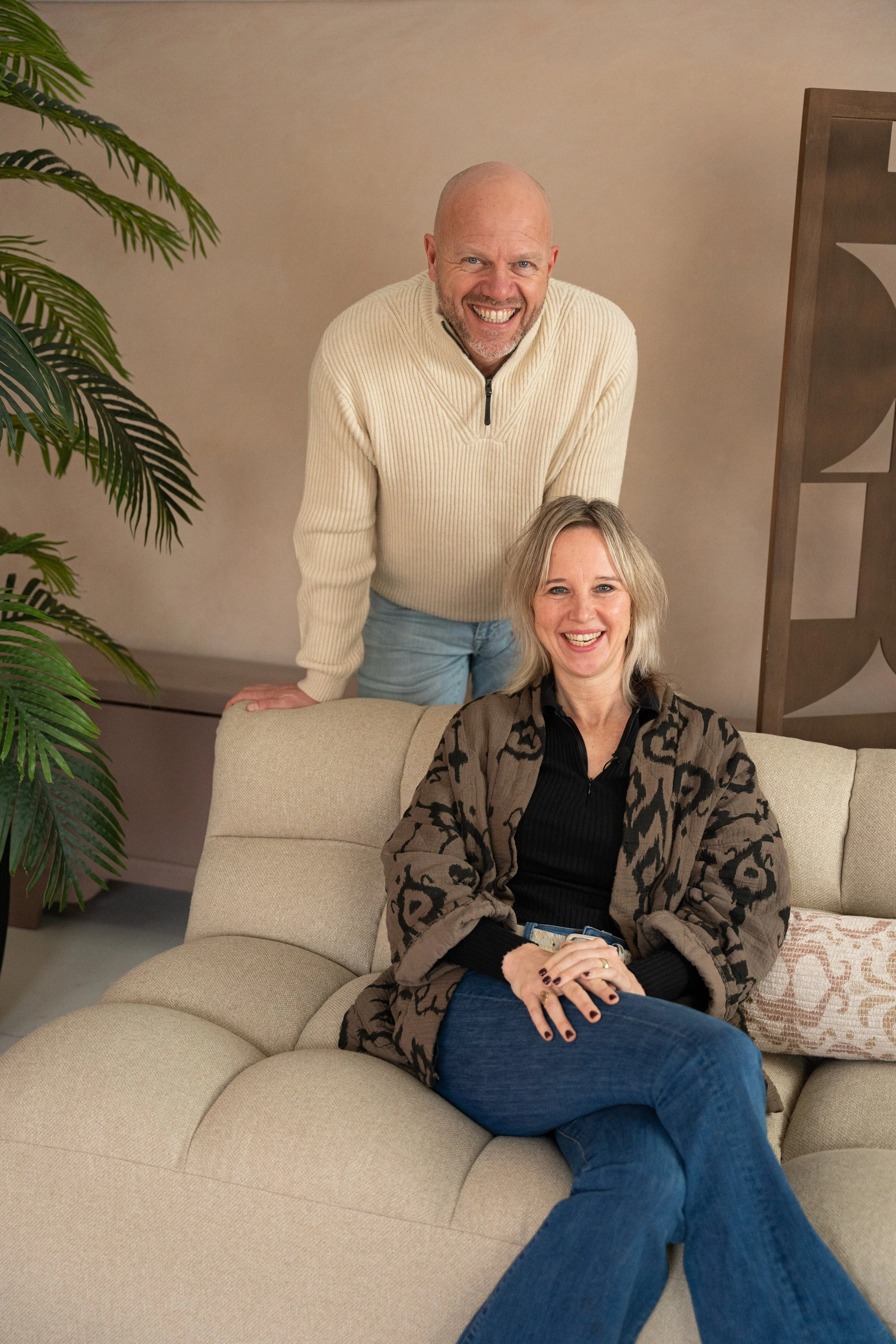 A man with a bald head and a goatee leaning on a beige sofa, smiling; a woman with shoulder-length blonde hair sitting on the sofa, also smiling, wearing a patterned cardigan and blue jeans; an indoor setting with a potted plant and decorative wooden panel in the background.