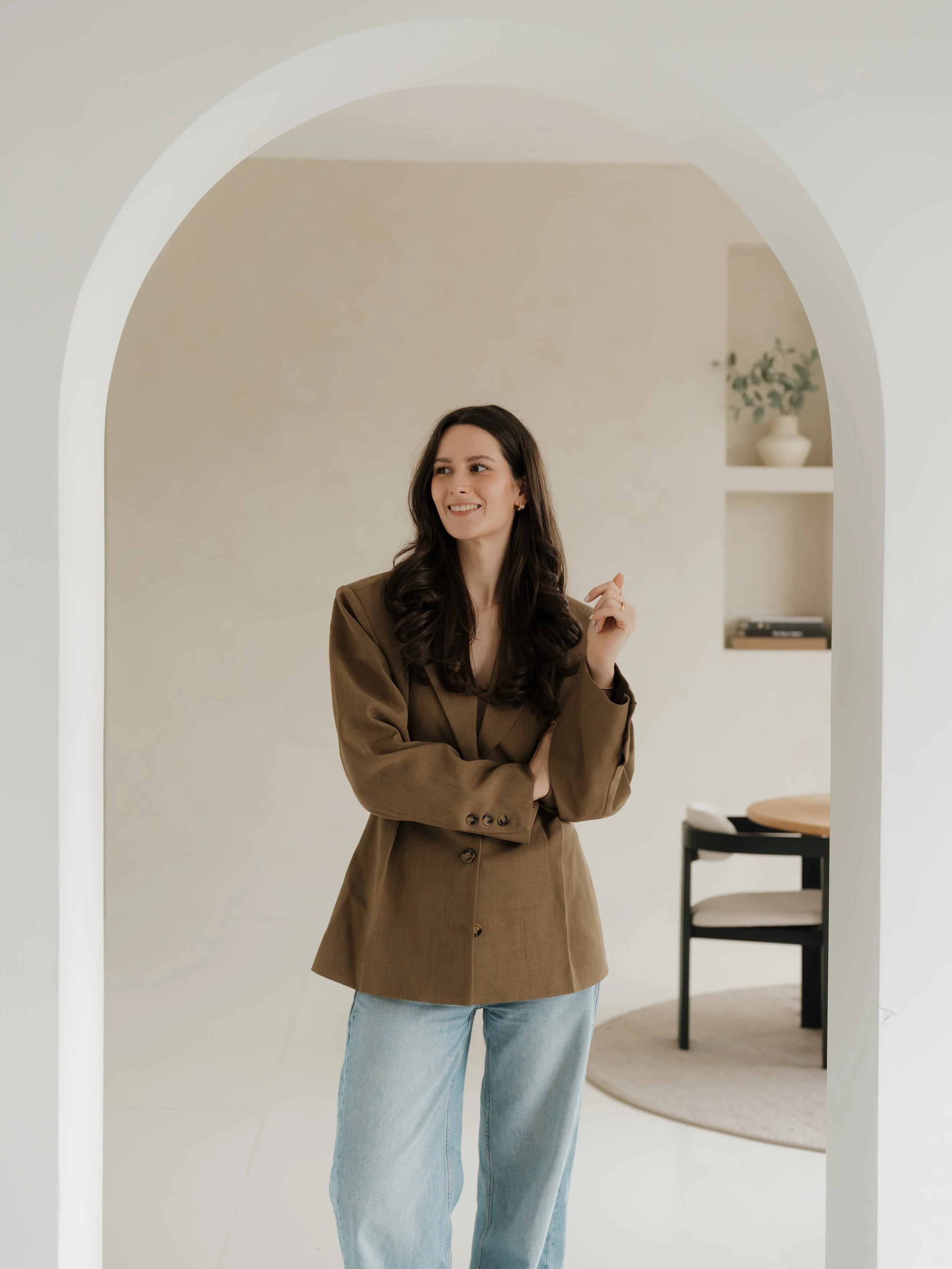 Woman with long dark hair smiling, wearing a brown blazer and light blue jeans, standing inside a modern home near an arched doorway.