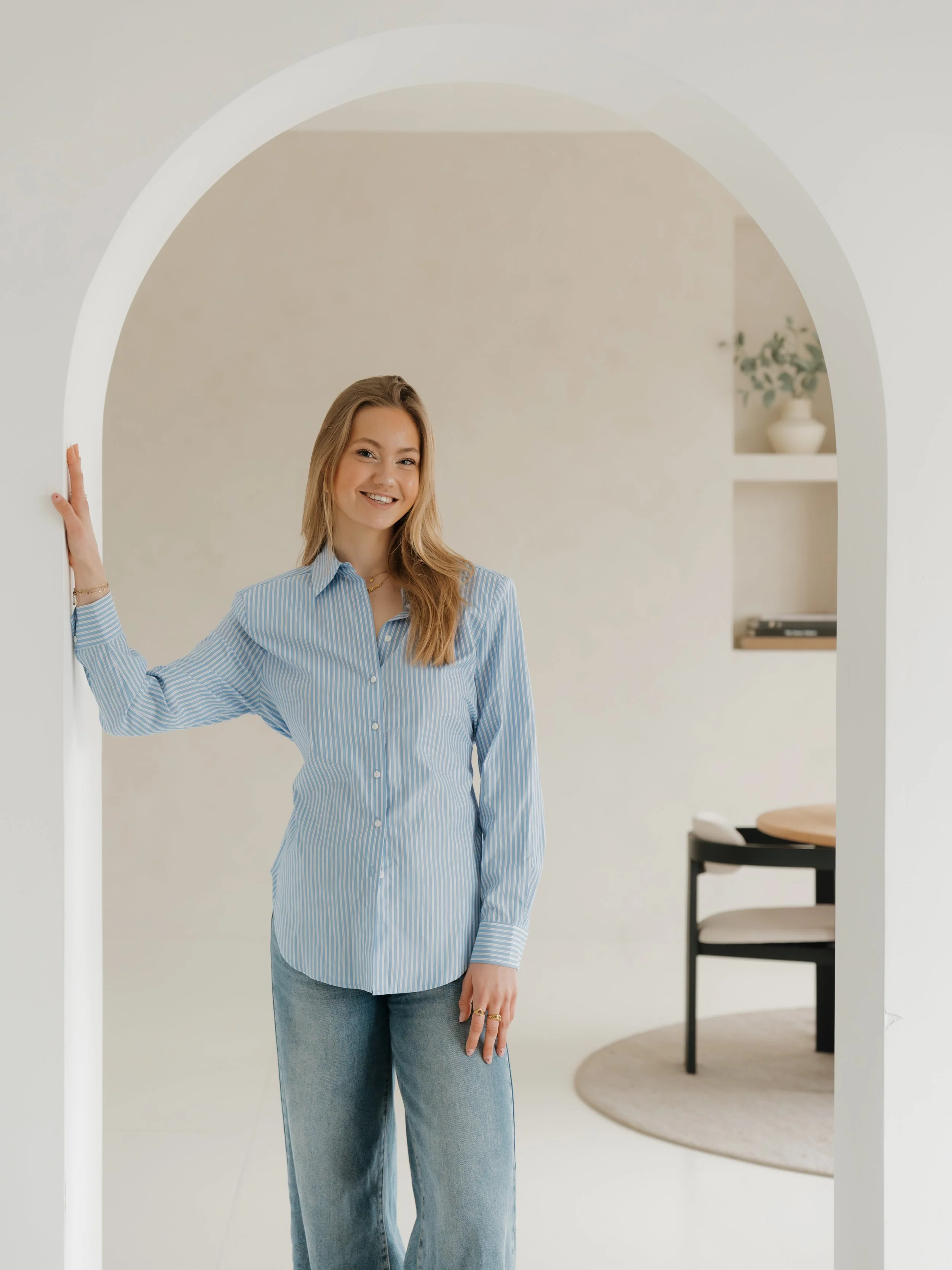 Young woman smiling, standing in doorway with arm resting on frame, wearing a blue and white striped shirt and jeans in a minimalist living space.