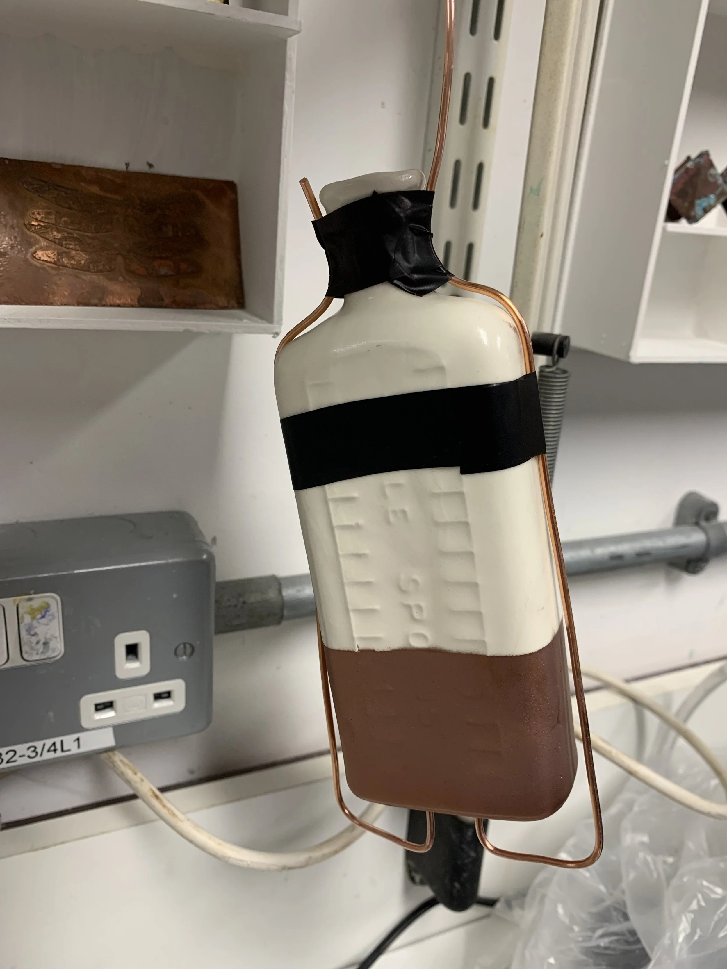 A makeshift device resembling a bottle with copper wiring and black tape, attached to a wall socket, possibly a DIY electronic project or repair.