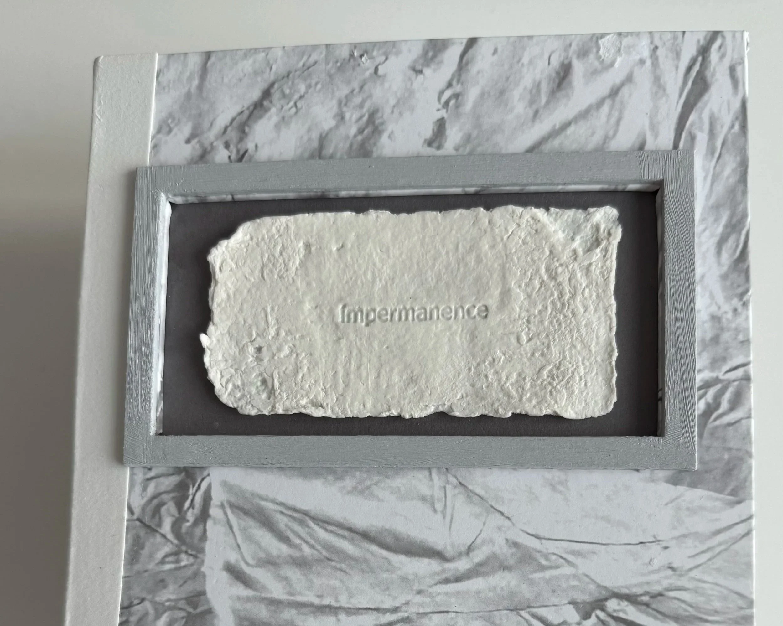 A hand holding a piece of white textured material with the word 'Impermanence' carved into it, mounted inside a grey wooden frame on an artist’s book.