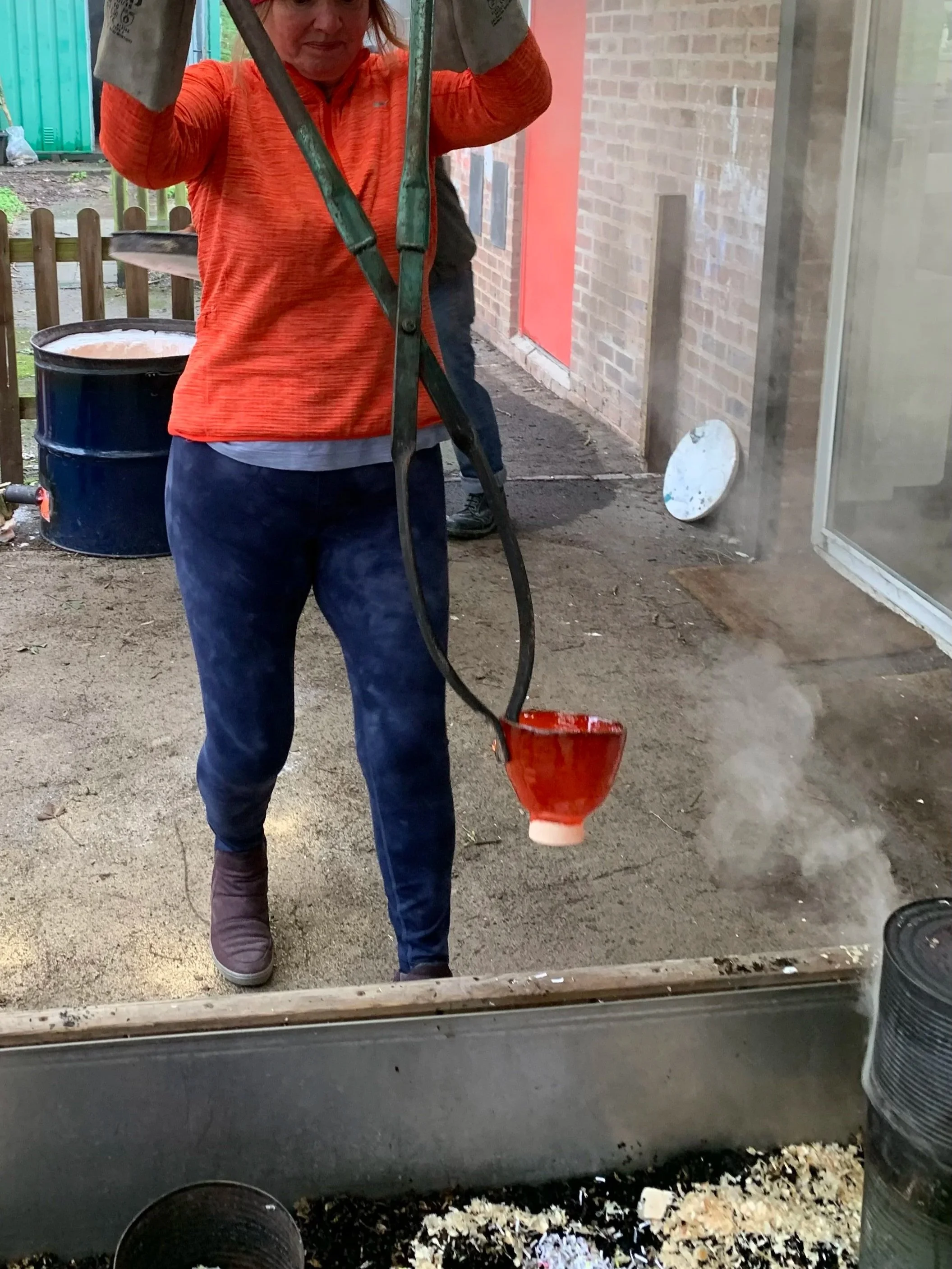 Woman in orange jacket and pink knit hat raku firing a piece of ceramic