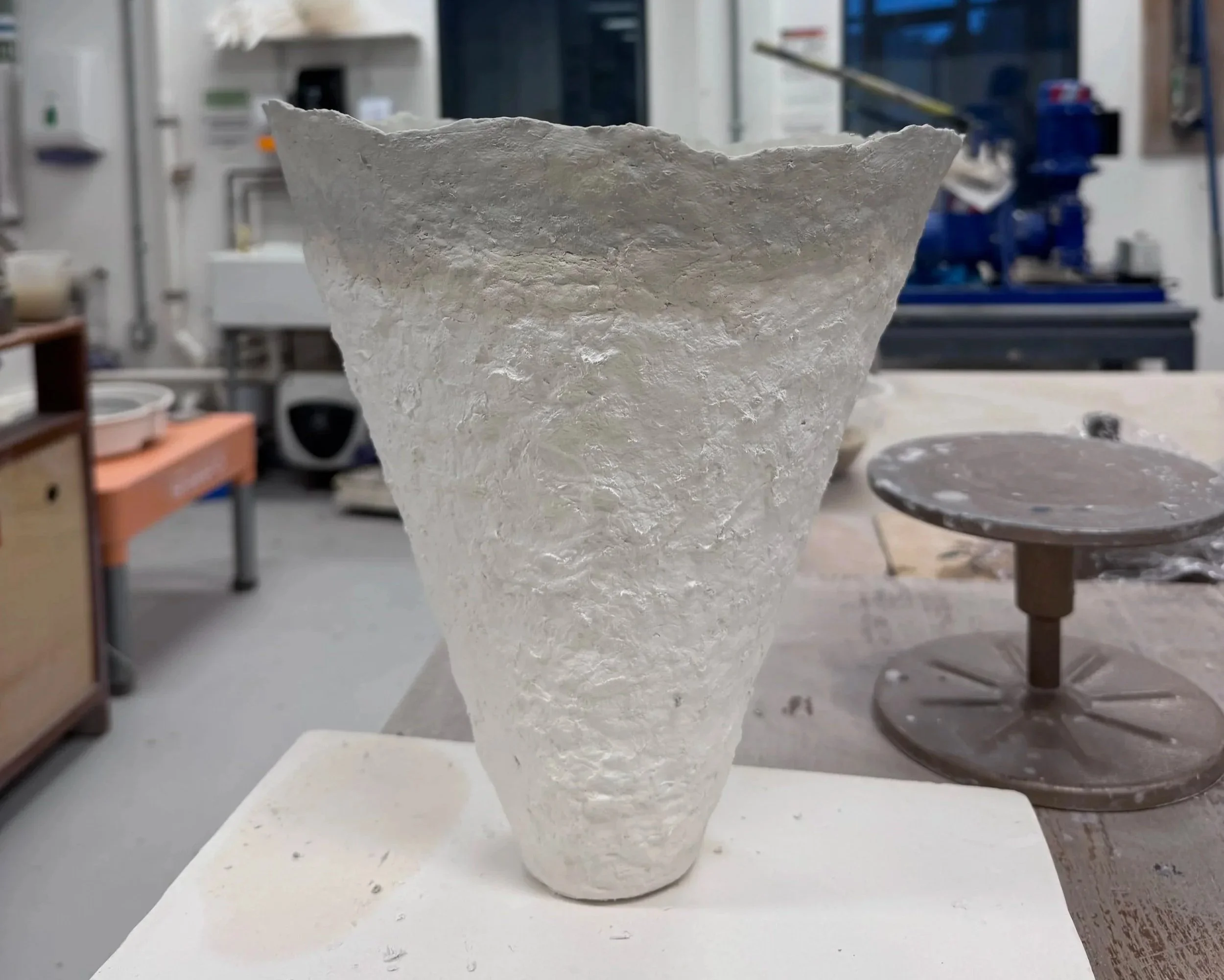 A textured, cone-shaped, white ceramic piece on a workspace with tools and equipment, displayed in an art studio or workshop.