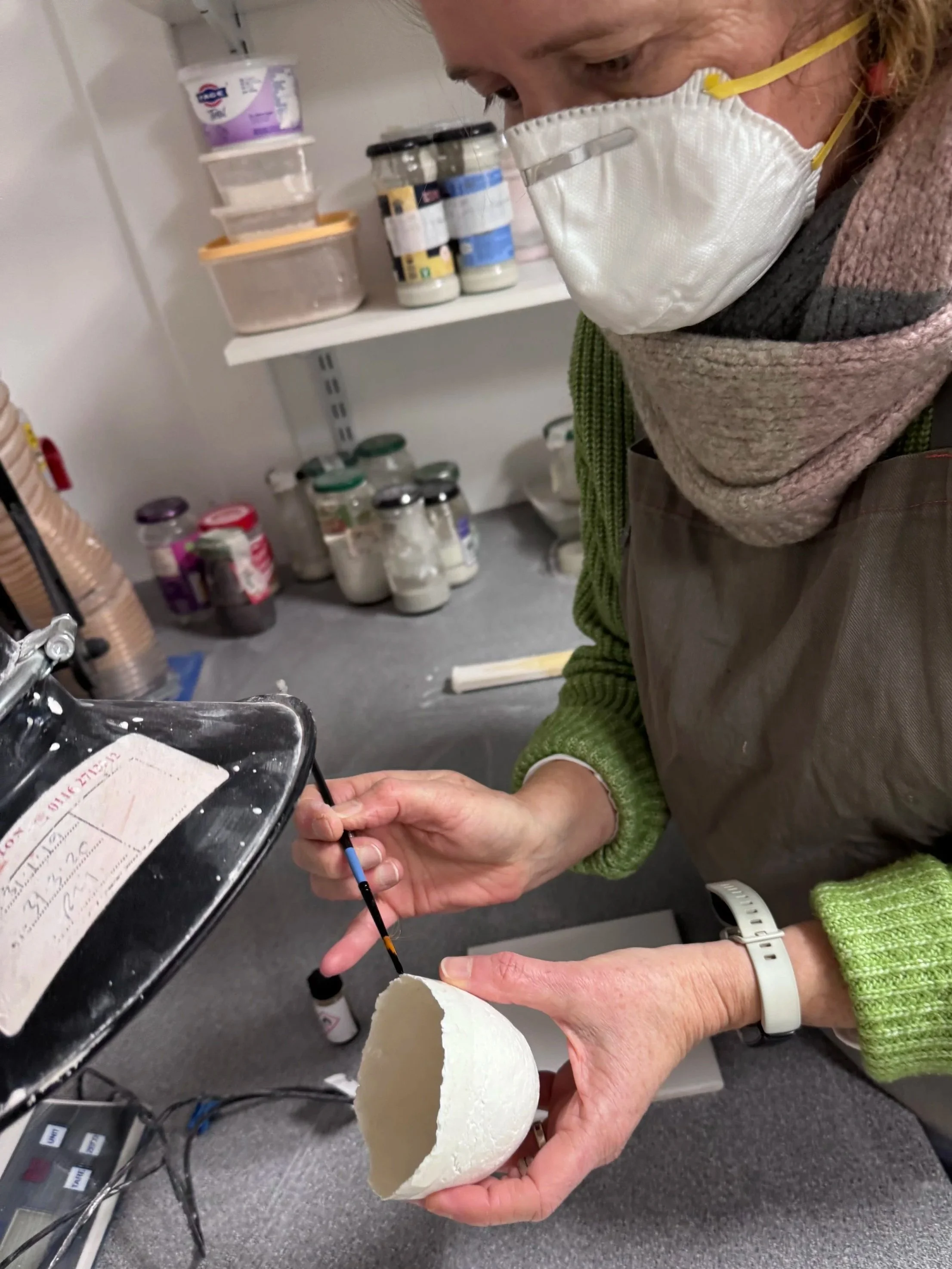 Person wearing a face mask and green sweater decorating a small white ceramic bowl with a black paintbrush in a laboratory or workshop setting.