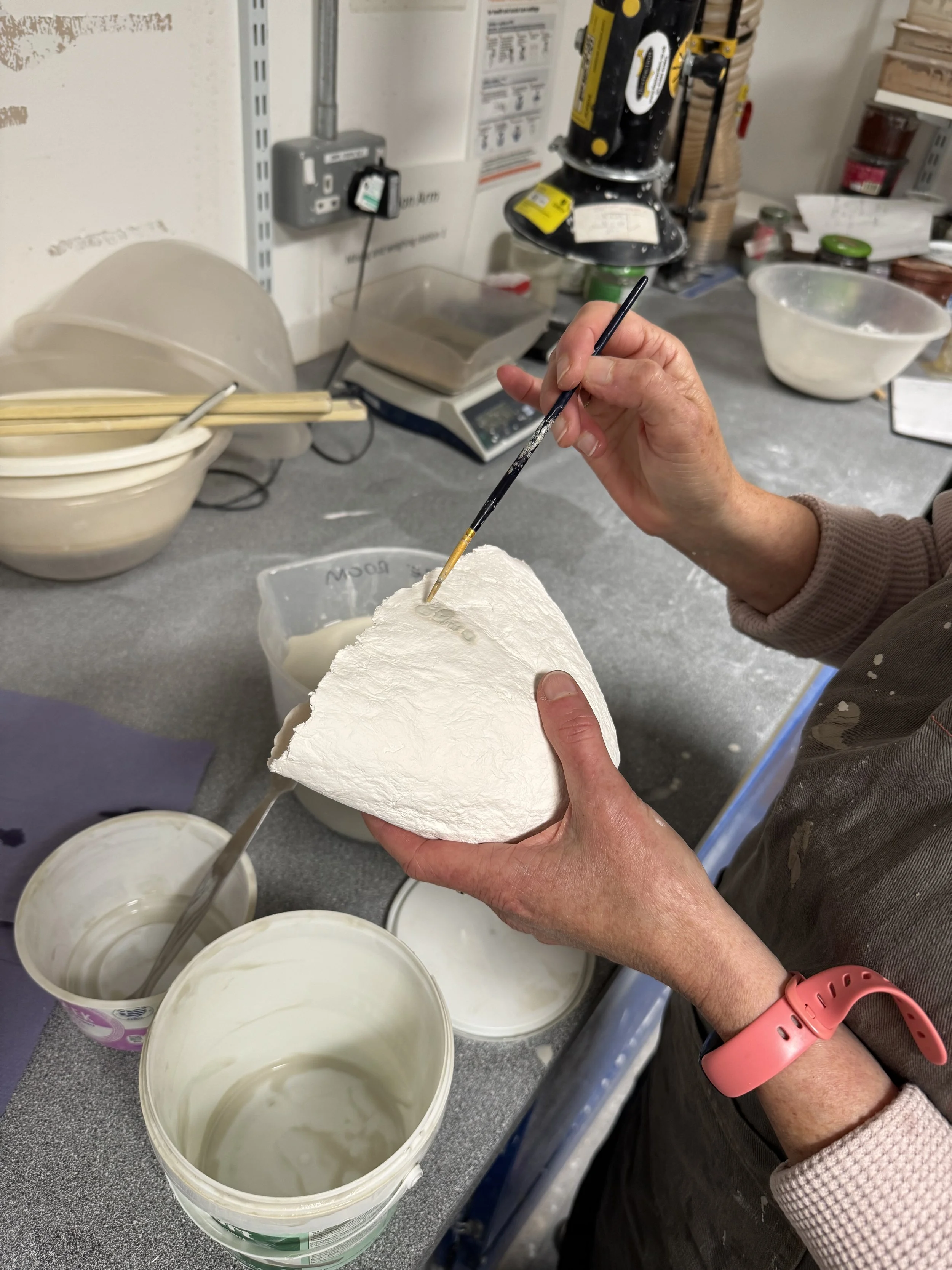 A person painting or decorating a large white surface, possibly made of plaster or ceramic, with a small brush in a workspace or studio. The workspace has bowls of paint or glaze, a scale, and various tools and supplies on the table.