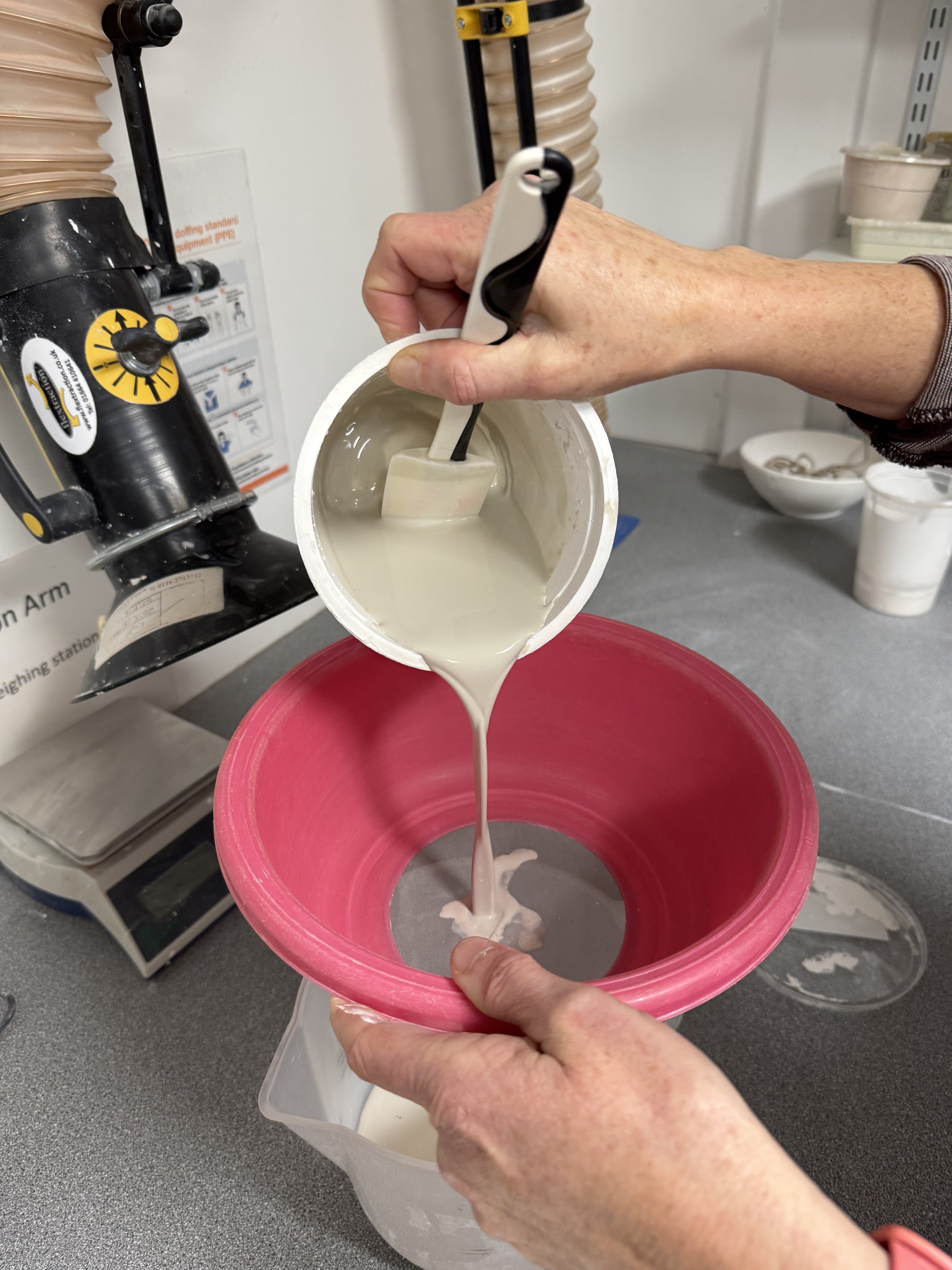 Person pouring a white liquid mixture through a sieve into a pink bowl, with a hand-held spatula submerged in the mixture.