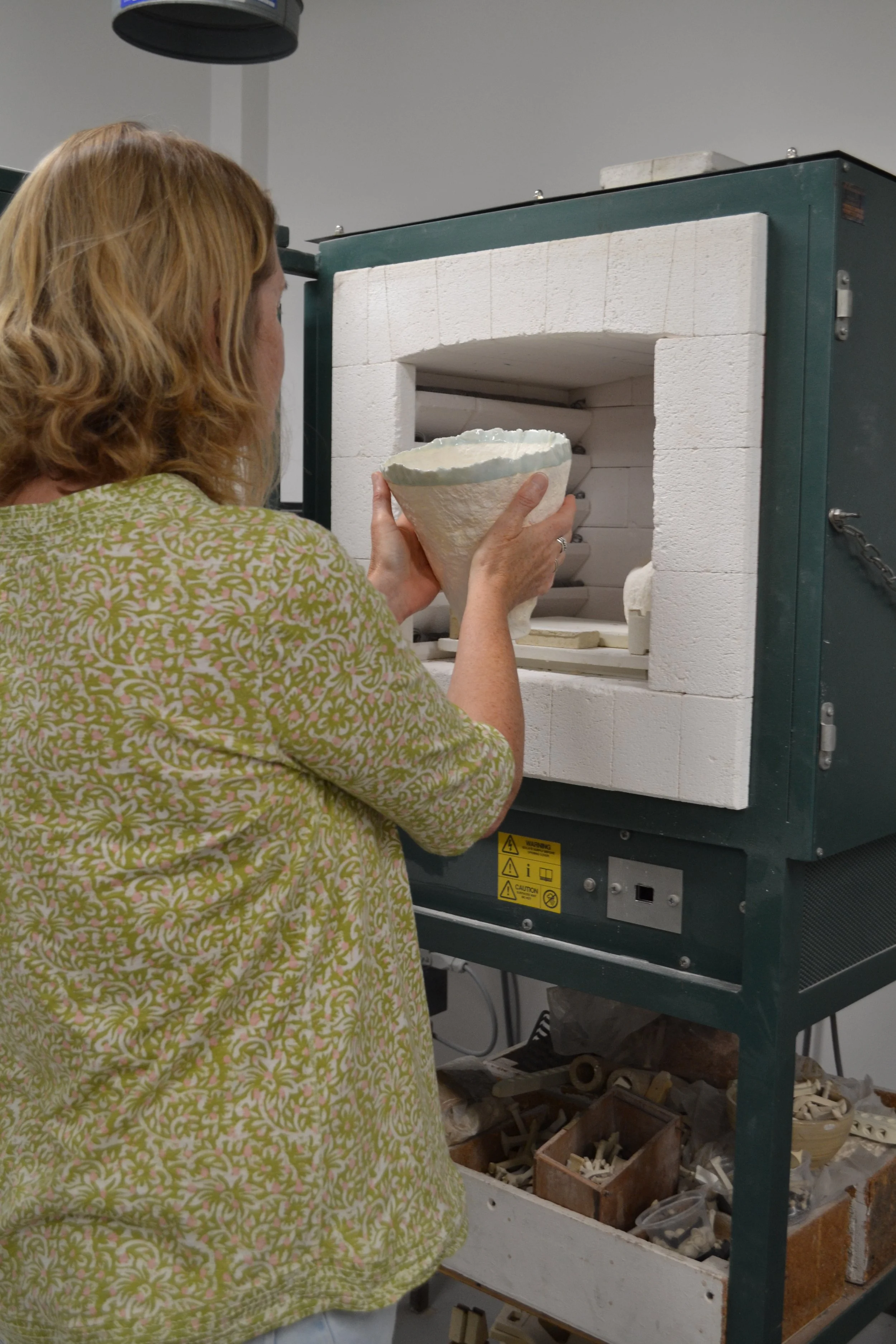 A woman in a patterned green top placing a ceramic piece into a kiln for firing.