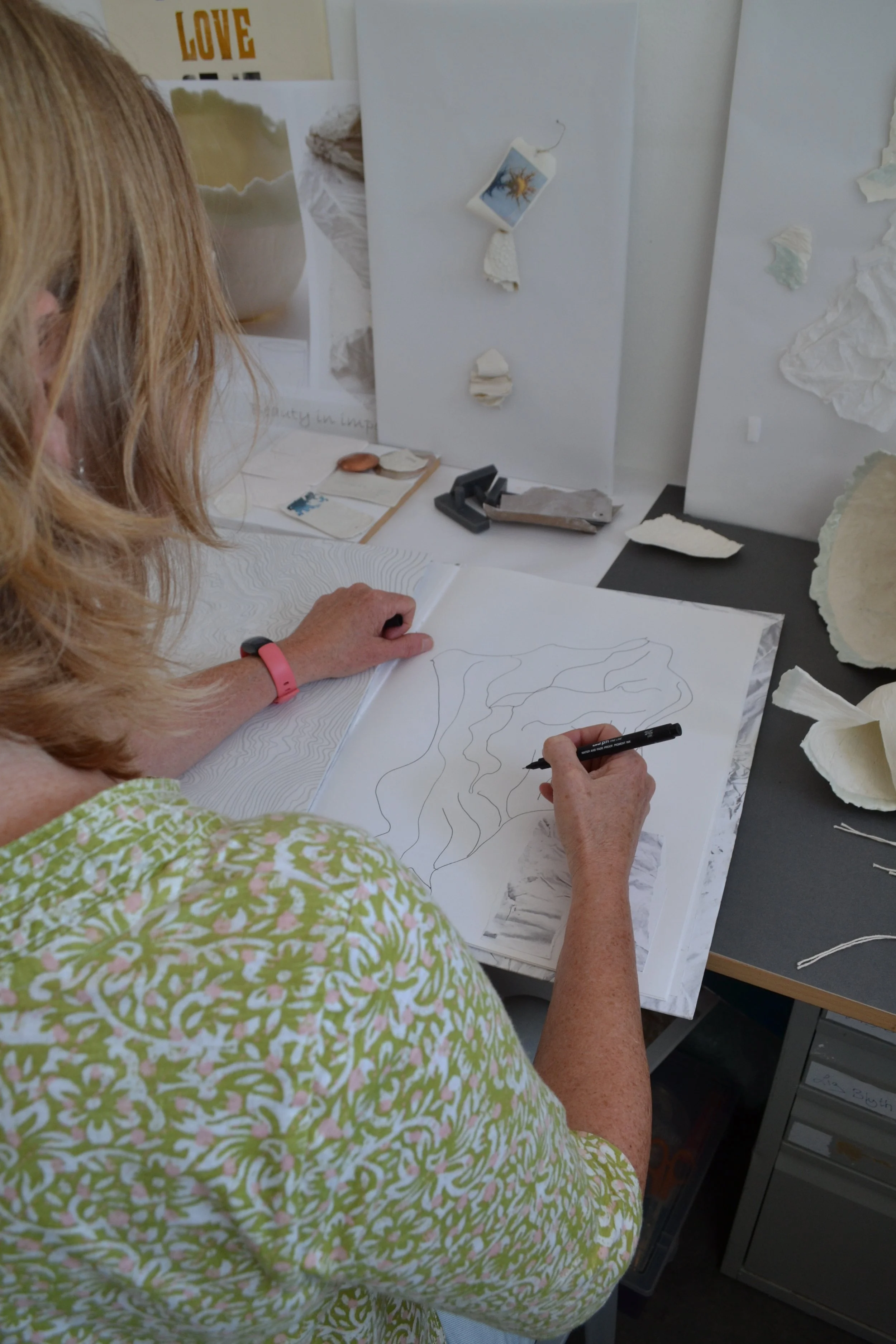A woman drawing a topographic map on a large sheet of paper at a workspace with art supplies and textured paper on the table.