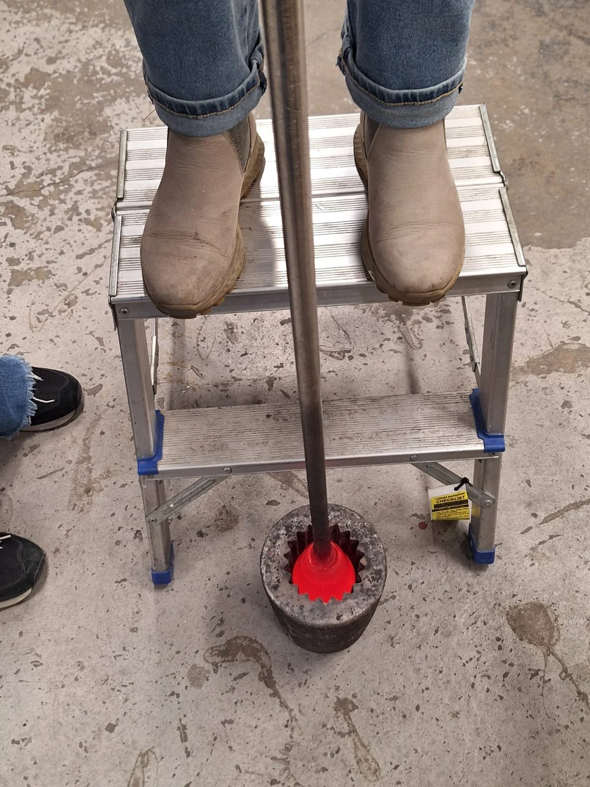A person standing on a small step ladder wearing beige work boots, with tools and a concrete floor visible.