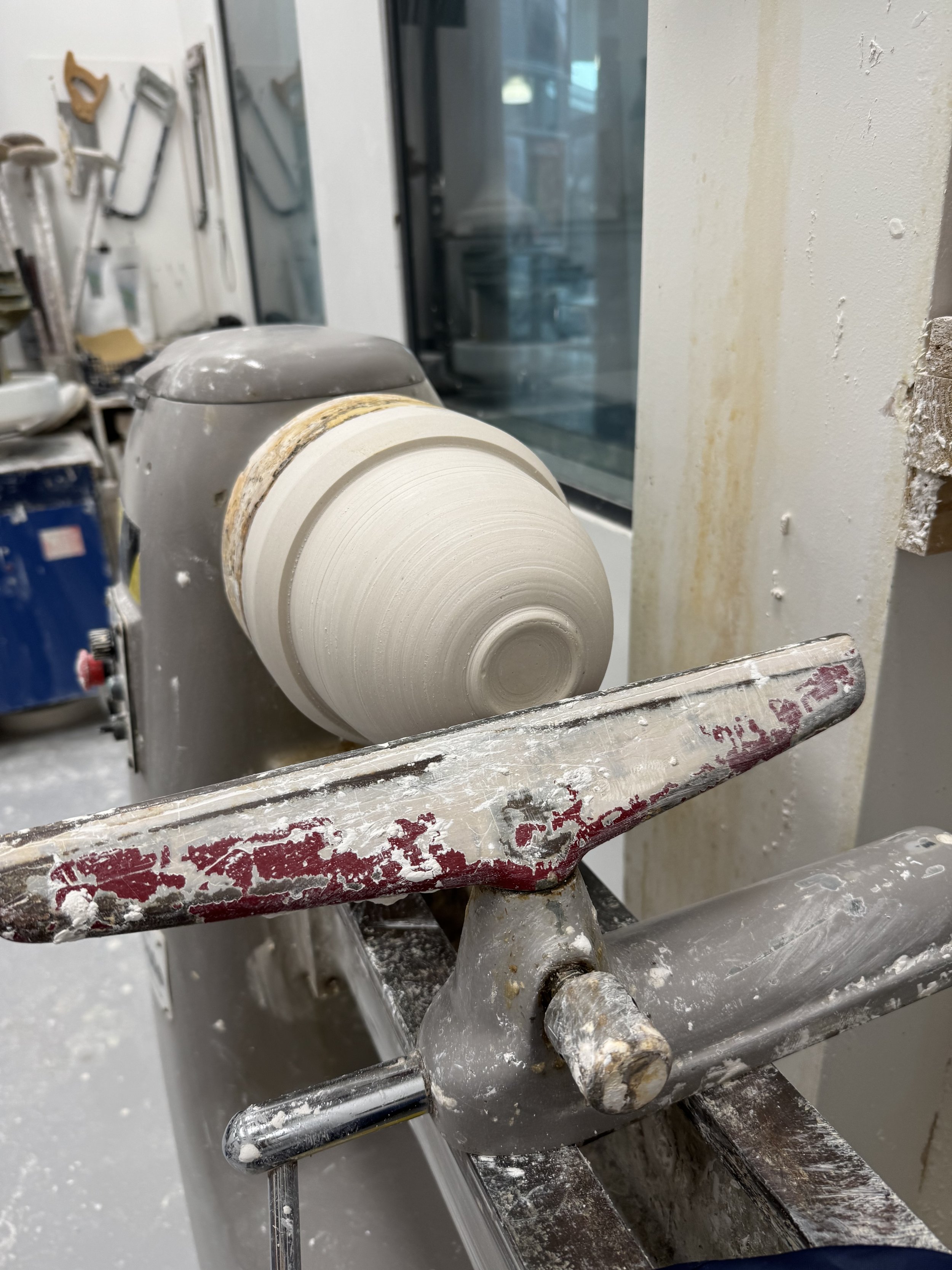 A pottery wheel in a workshop with a spinning clay piece mounted on it, surrounded by dust and pottery tools in the background.
