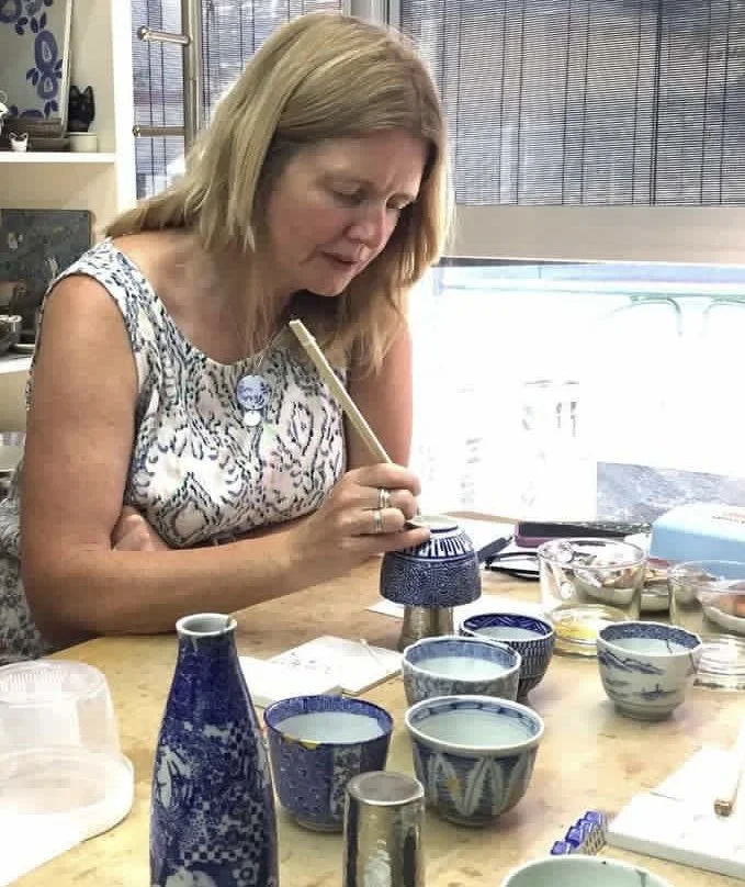 A woman decorating ceramic items with a brush at a table in a well-lit room.