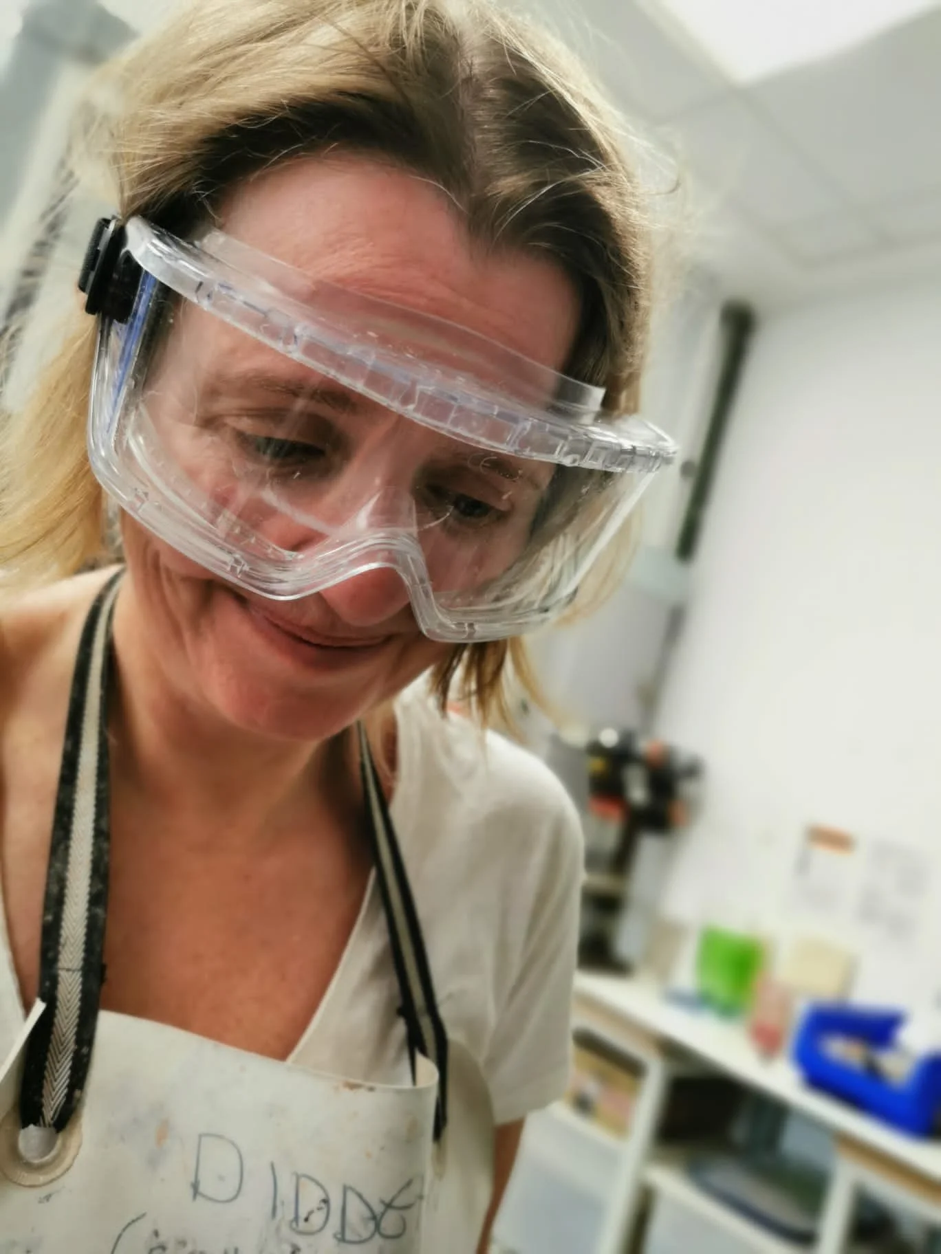 A woman wearing safety goggles and a white apron in a workspace, smiling and looking down.