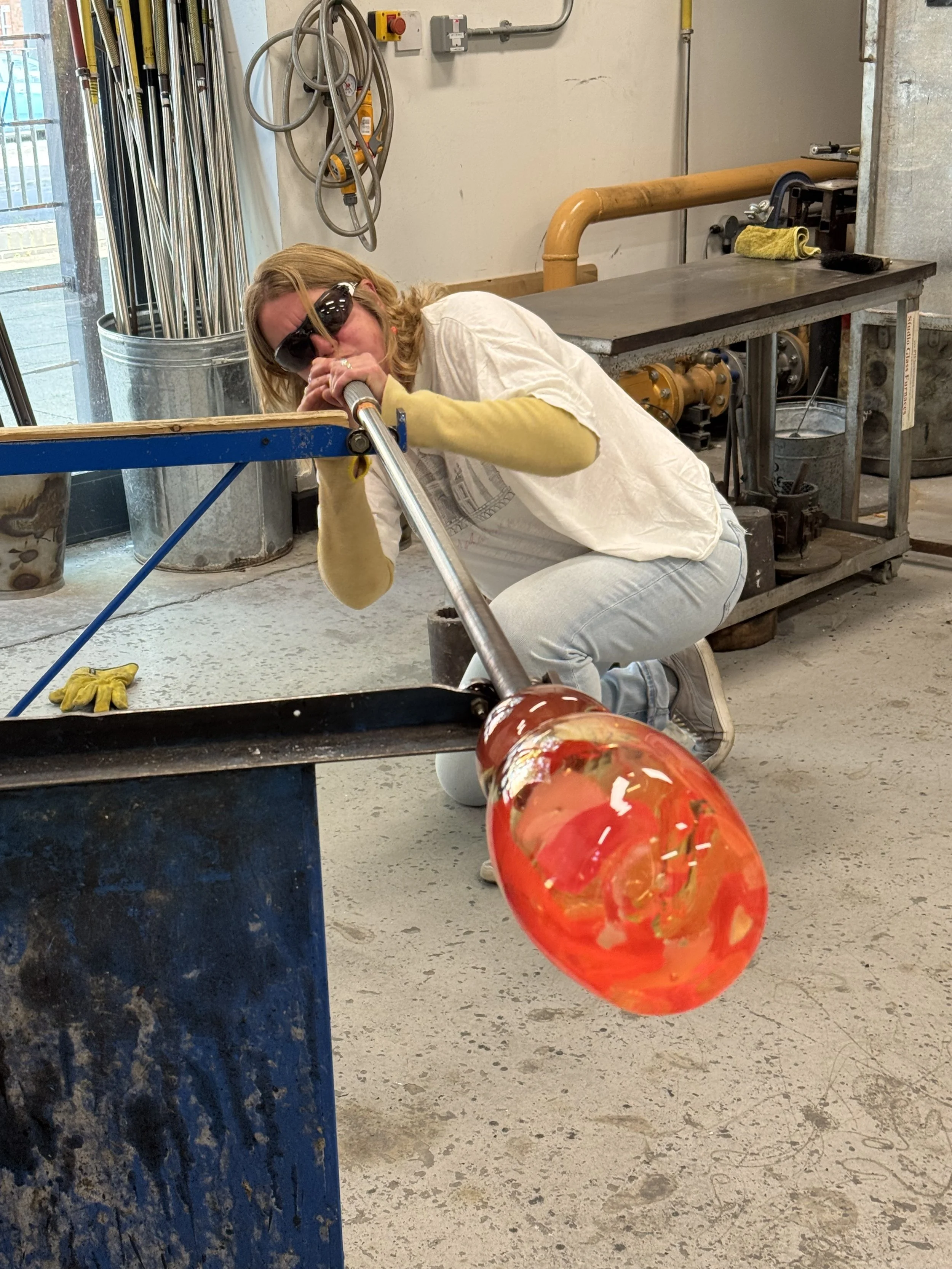 A woman glassblowing a red and orange glass piece in a workshop, kneeling on the floor and blowing hot glass