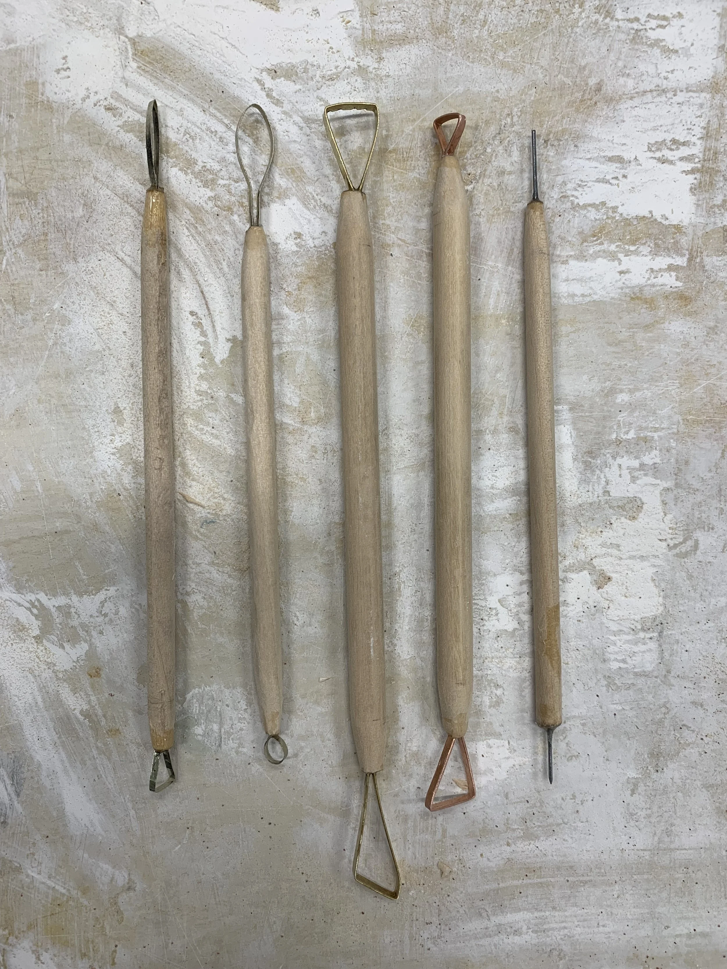 Six sculpting tools with wooden handles and various metal tips, laid out horizontally on a textured surface.