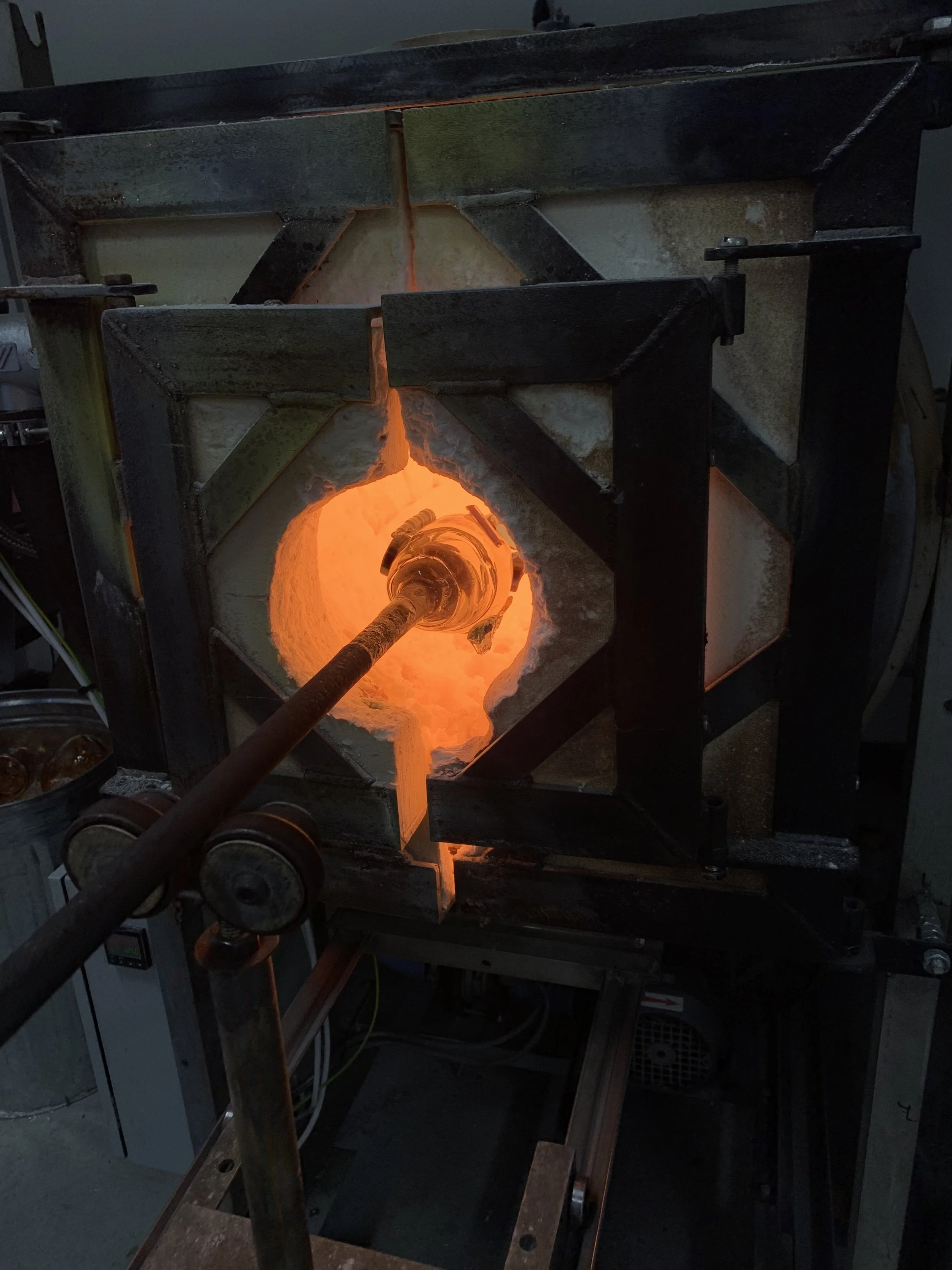 A glassblowing furnace with a glowing orange-hot glass piece inside, surrounded by a metal framework.