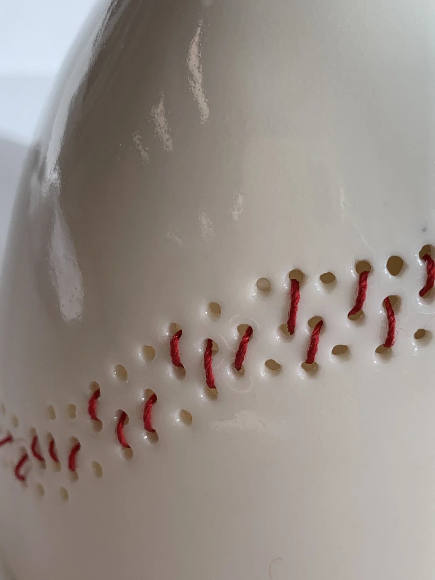 Close-up of a white glazed piece of ceramic with small holes and red thread passing through some of the holes, forming a pattern.