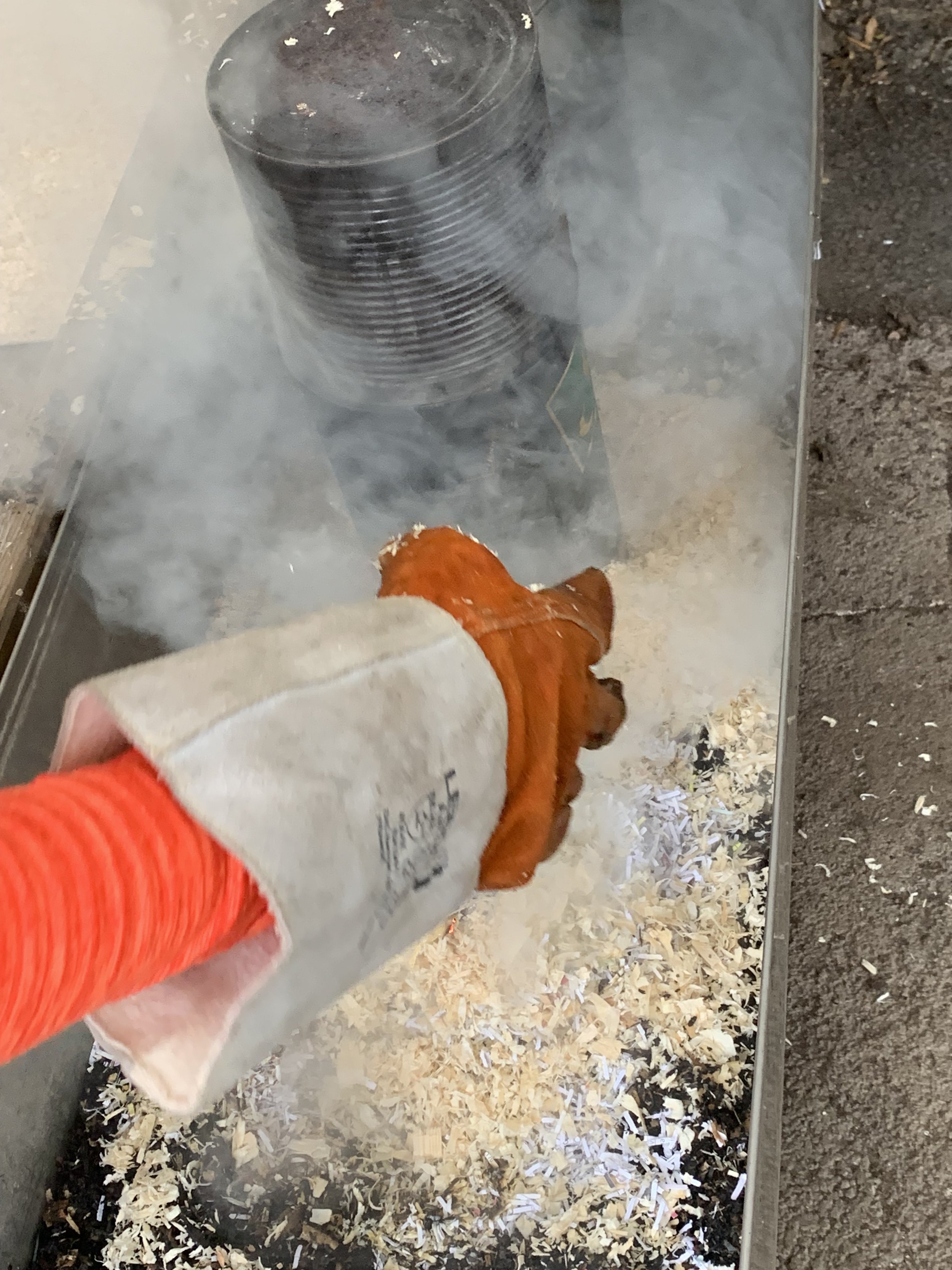 A person wearing a heat proof work glove is placing a ceramic vessel in a pit of sawdust and wood shavings.