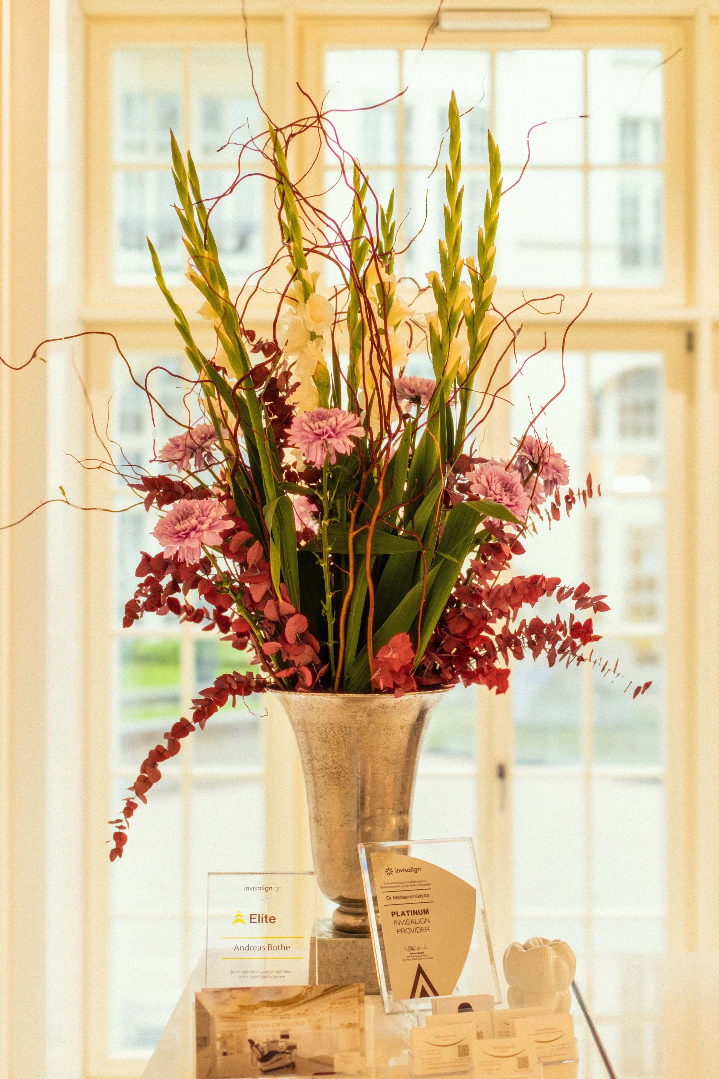 Blumenvase mit weißen und rosa Blumen auf einem Tisch vor einem großen Fenster in einer hellen Zahnarztpraxisumgebung.