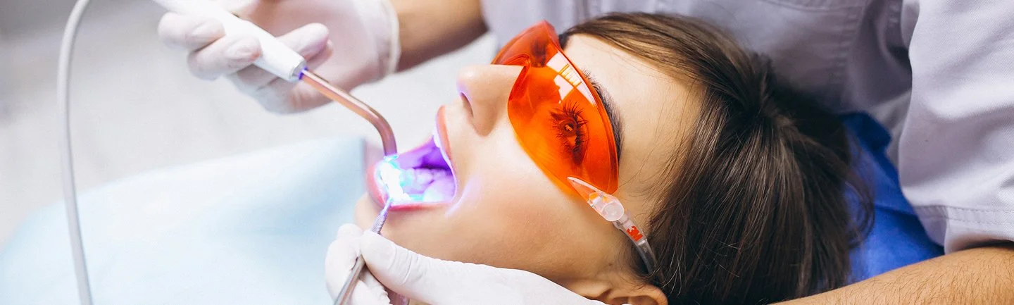 Patientin liegt im Behandlungsstuhl und erhält fotoaktive Lasertherapie, Zahnarzt behandelt die Zähne mit UV-Licht, Schutzbrille getragen