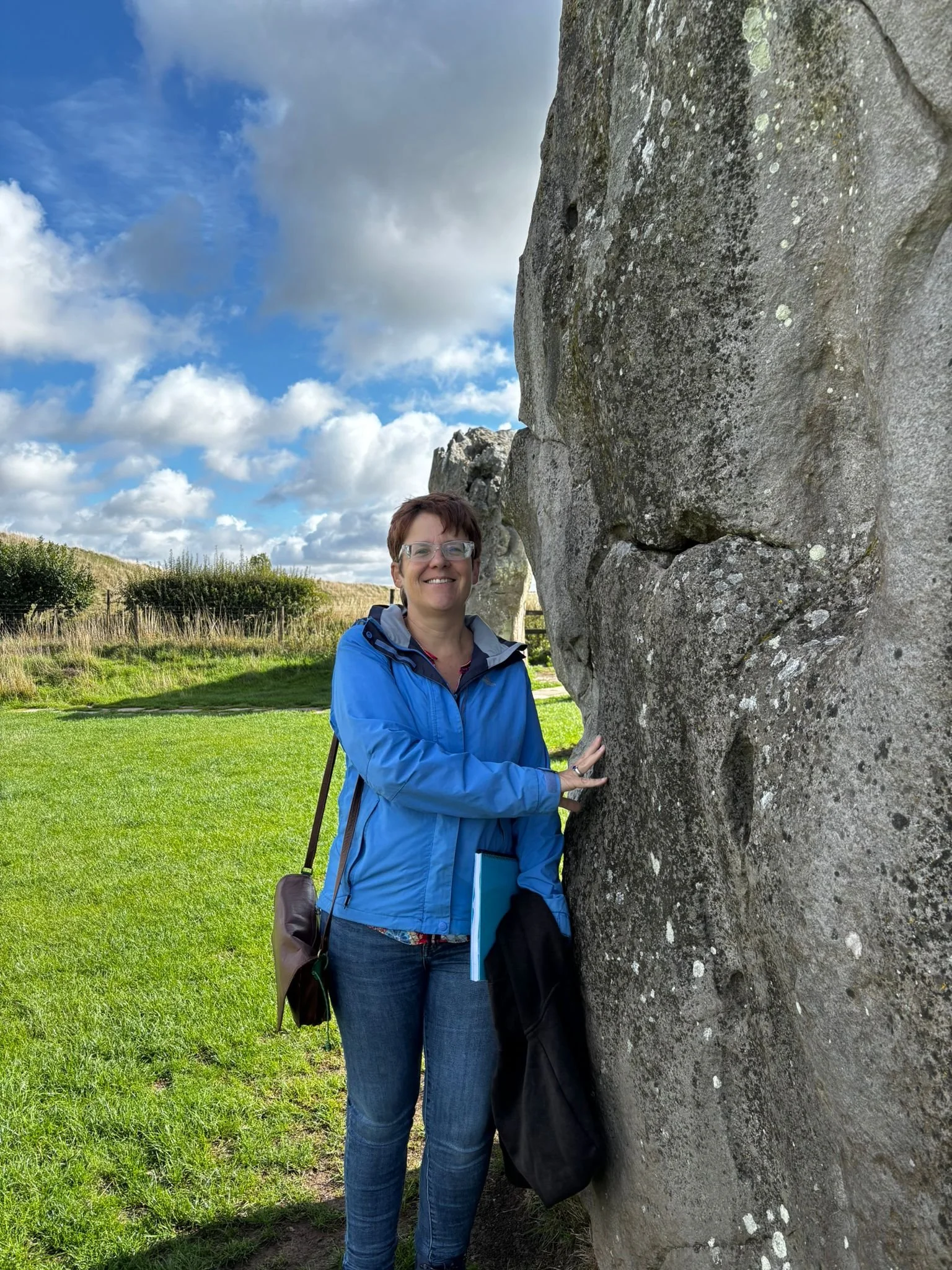 Helen at Avebury touching one of the standing stones wearing a blue anorak on a cloudy day