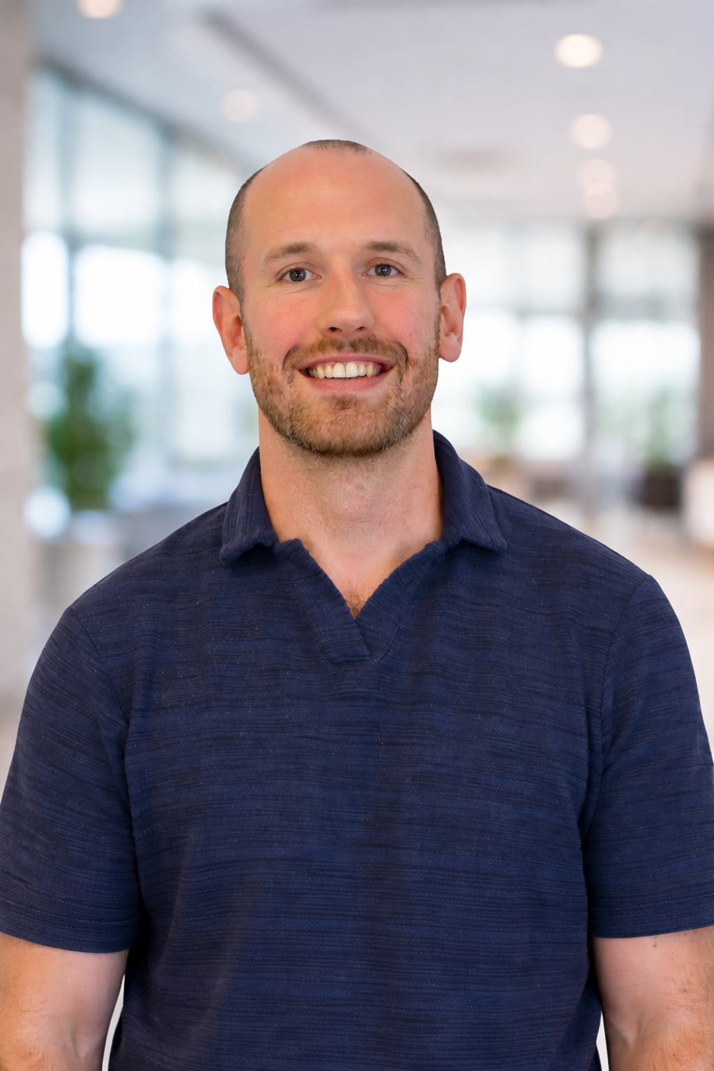 A smiling man with a beard and short hair wearing a navy blue polo shirt in a bright, modern office space with large windows and blurred background.