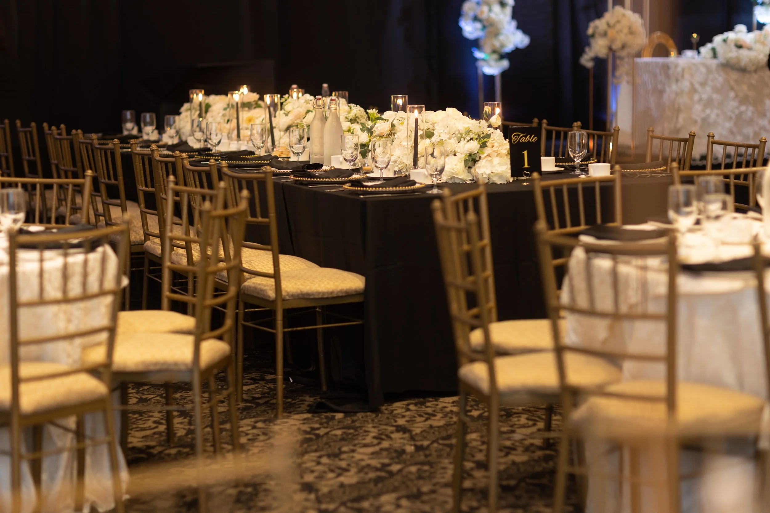 Elegant banquet table setup with white floral centerpiece, candles, and gold chairs in a dimly lit room for a formal event.