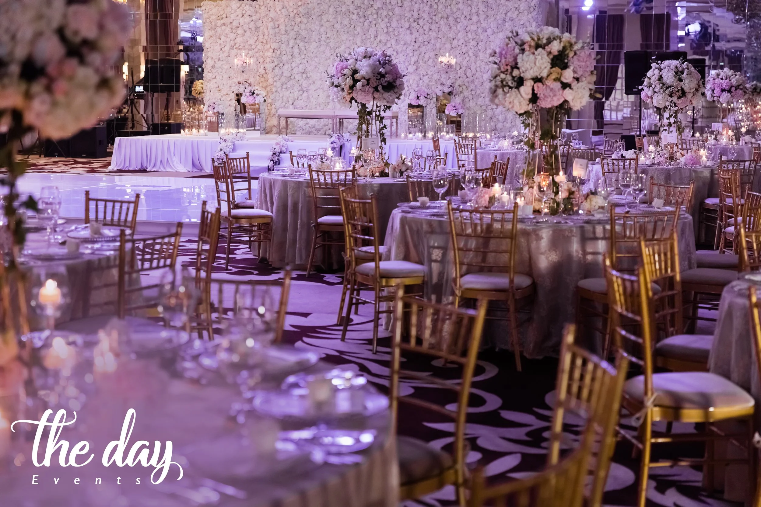 Elegant banquet hall decorated with large floral centerpieces, tables with candles, and gold chairs, prepared for a wedding reception or event.