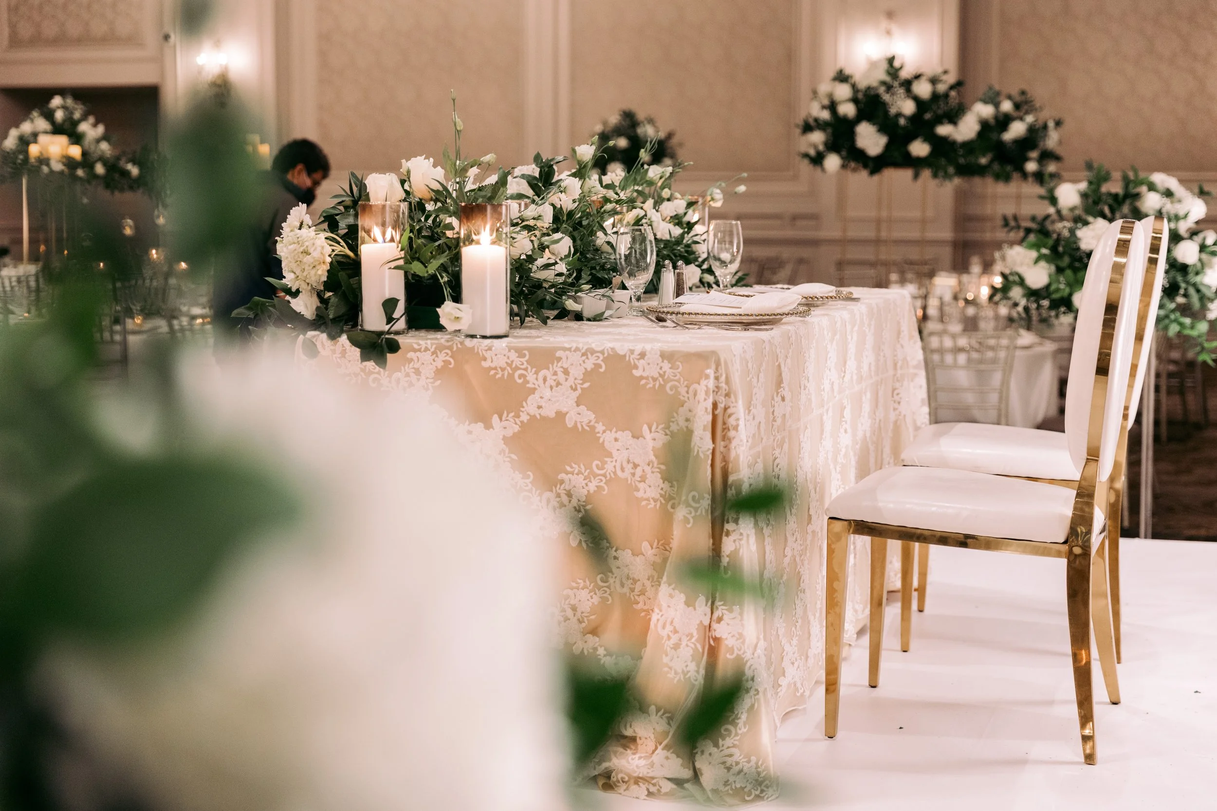 Elegant banquet table decorated with white and green floral arrangements, tall candles, glassware, and gold-trimmed chairs, set in a luxurious room with ornate wall details.