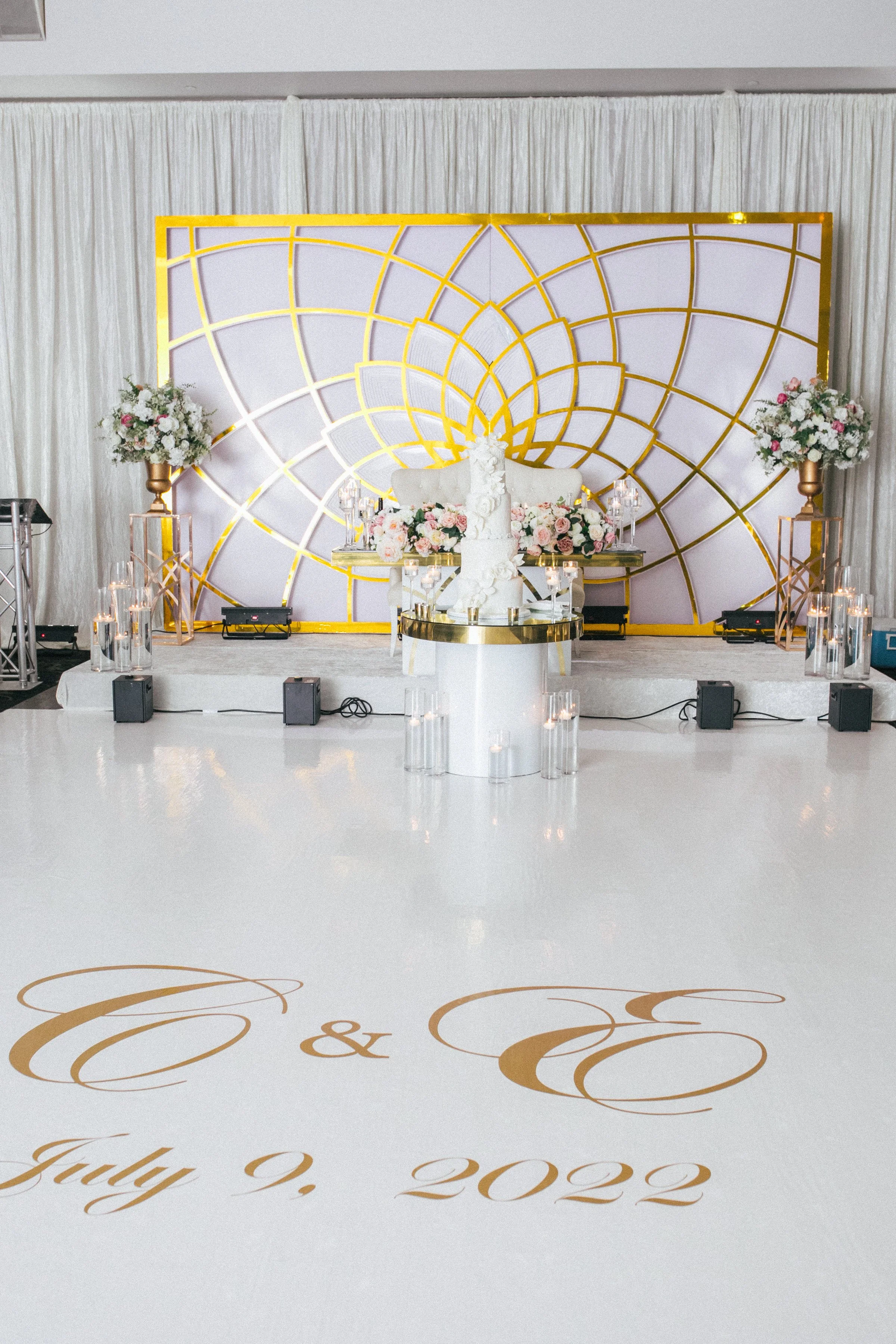 Wedding reception setup with a white dance floor, gold accents, and floral arrangements. Gold geometric backdrop behind a table with a wedding cake. The dance floor has gold calligraphy with the initials "C & E" and the date "July 9, 2022."