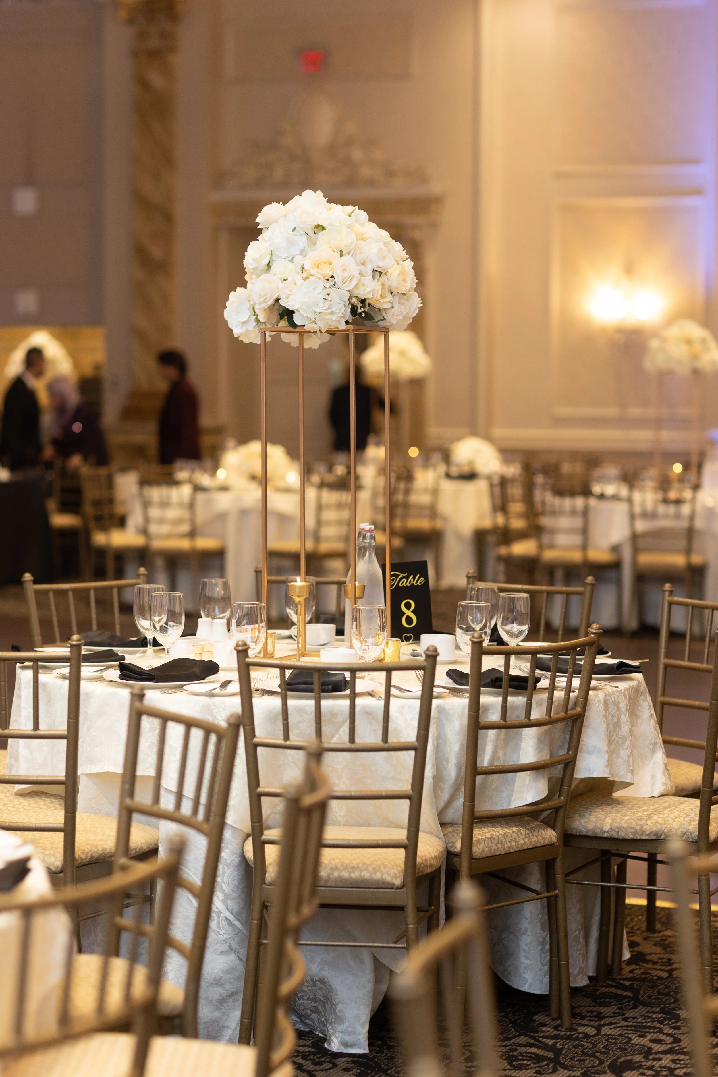 Elegant banquet table setup with a white floral centerpiece on a tall gold stand, surrounded by gold chairs and tableware in a decorated event hall.