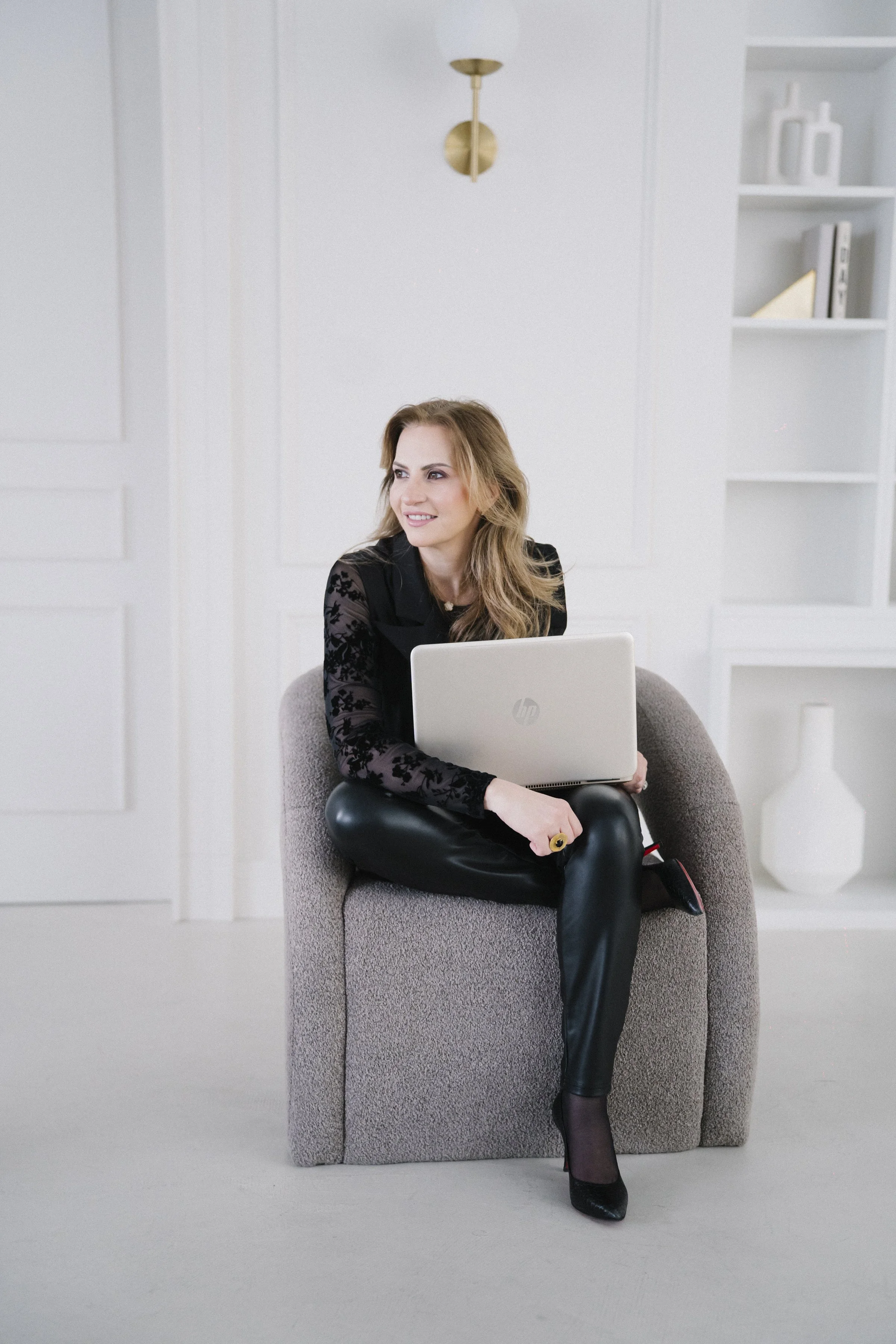 A woman sitting on a beige armchair with a laptop on her lap in a bright, modern room with white walls and a bookshelf.