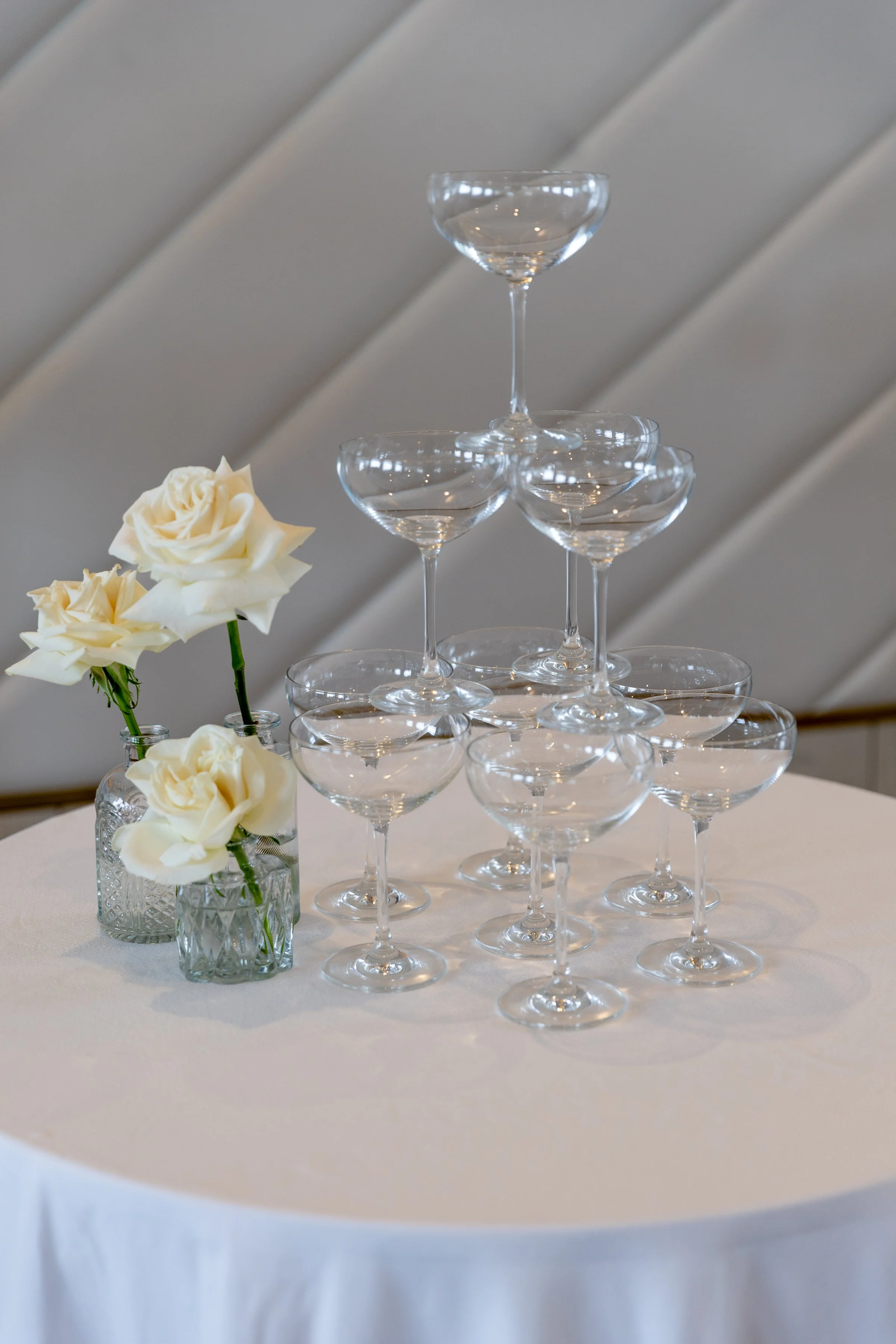 A pyramid of empty champagne glasses on a table with white tablecloth, alongside two small vases of white roses.