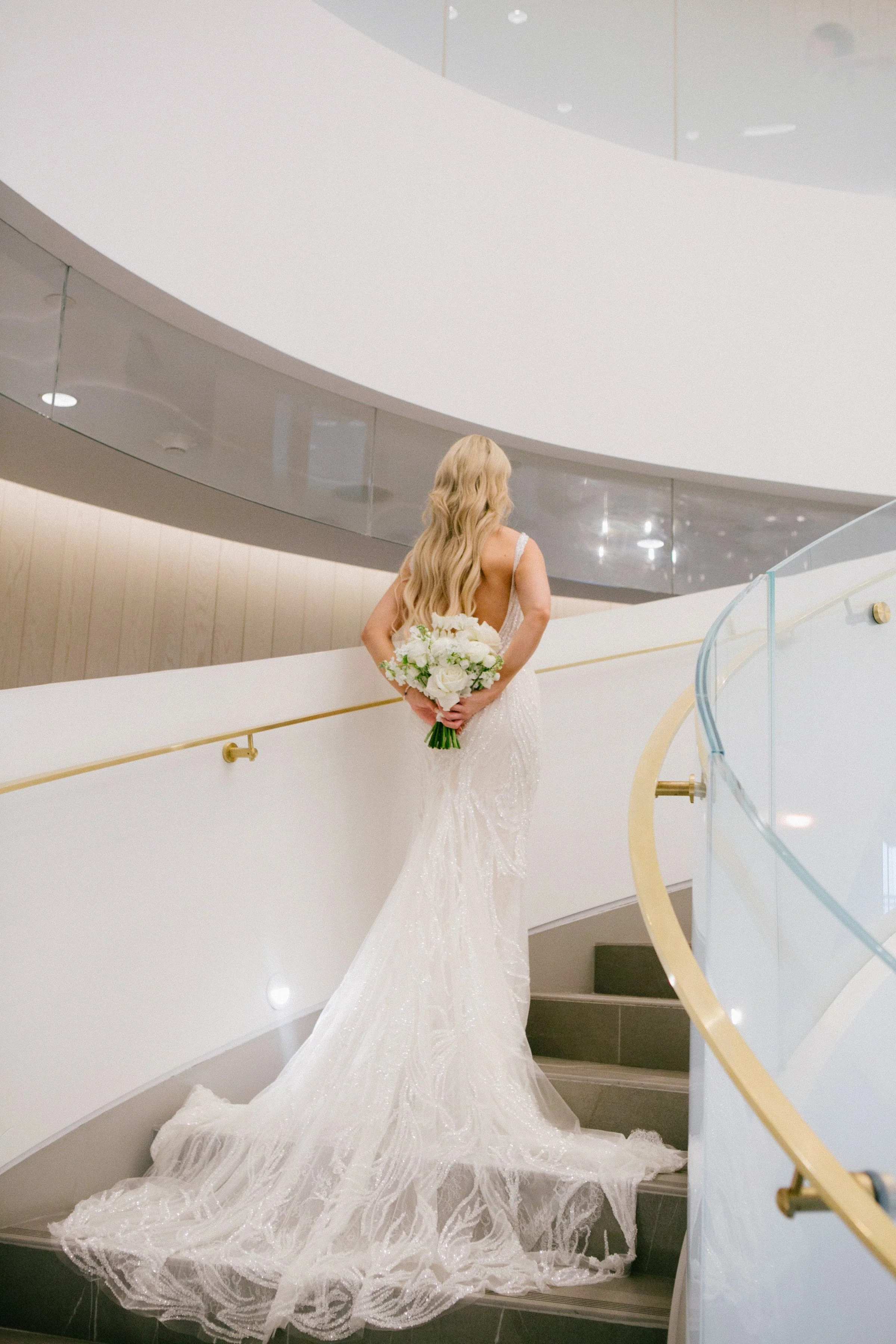 A bride in a white wedding gown holding a bouquet of white flowers on a modern staircase.