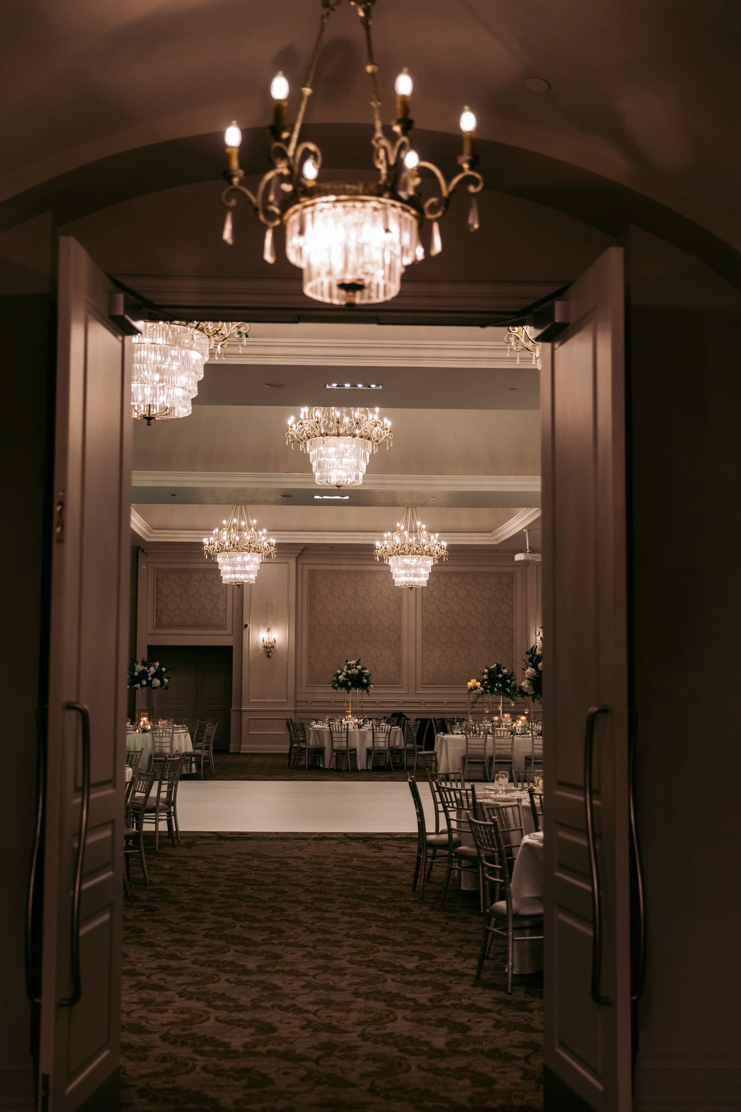 Elegant banquet hall with chandeliers, decorated round tables, and floral centerpieces, viewed through open double doors.