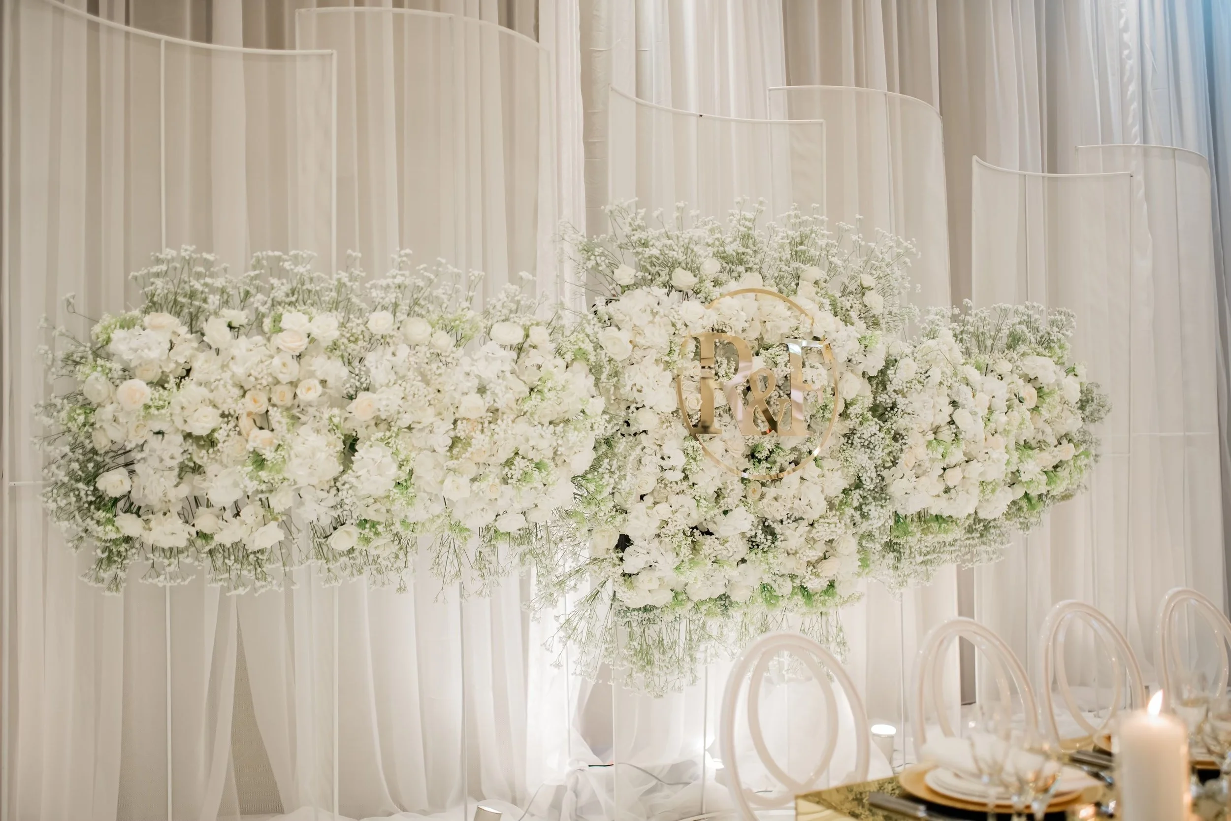 White floral arrangement with a monogrammed gold 'R & F' in the center, set against a backdrop of white drapes and decorated tables with white chairs and candles.