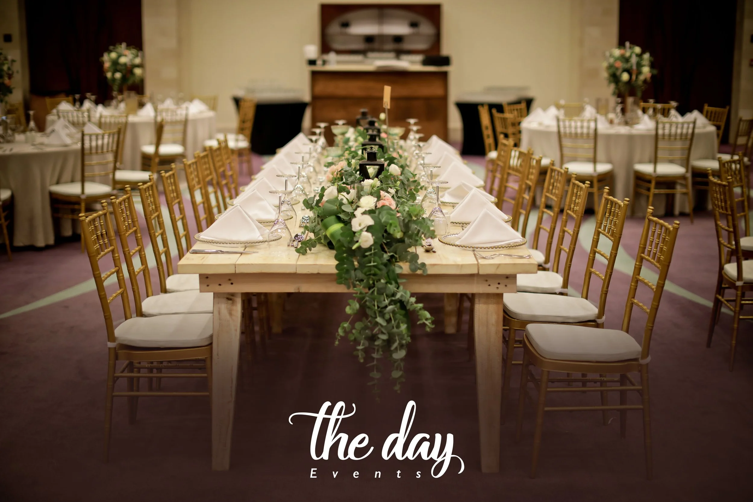 A decorated banquet table with white napkins, glassware, and a green floral centerpiece, set in an event hall with other round tables, gold chairs, and a wooden piano in the background.