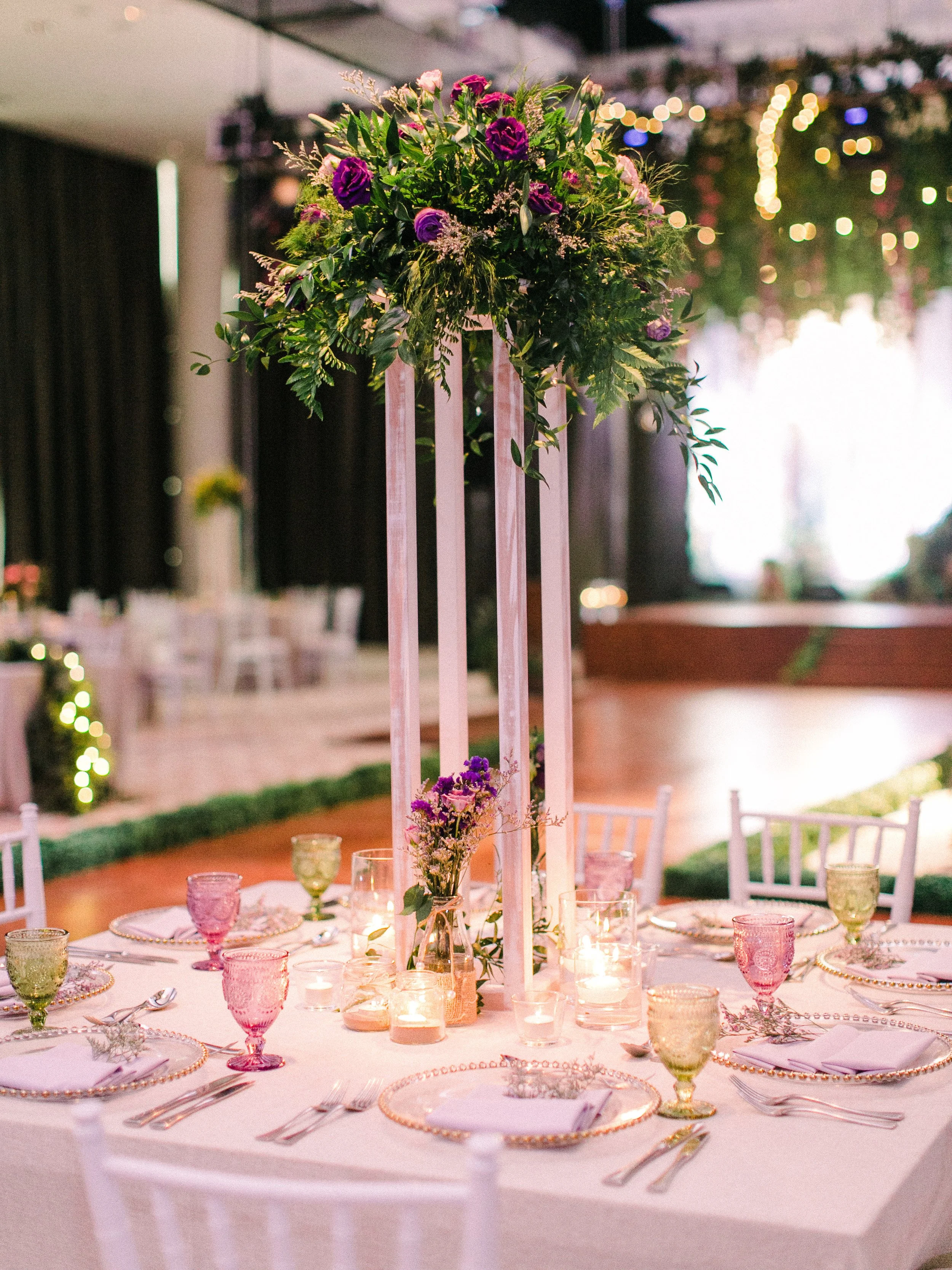 Decorated round banquet table with center floral arrangement, candles, pink and green glasses, and place settings in an event space with string lights.