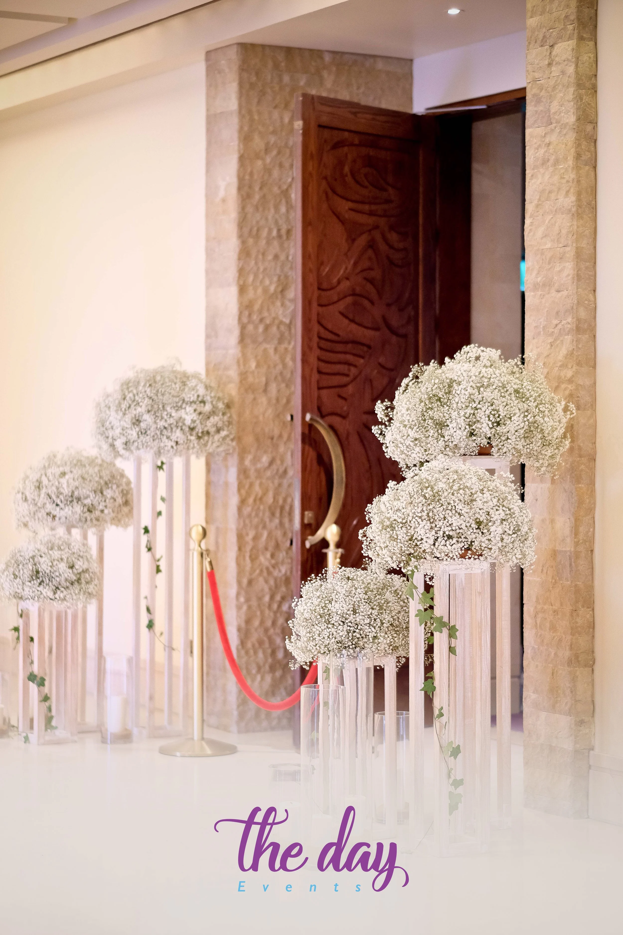 Elegant event entrance with white baby's breath floral arrangements on tall clear vases, a red rope barrier, and a wooden door with carved patterns in the background.