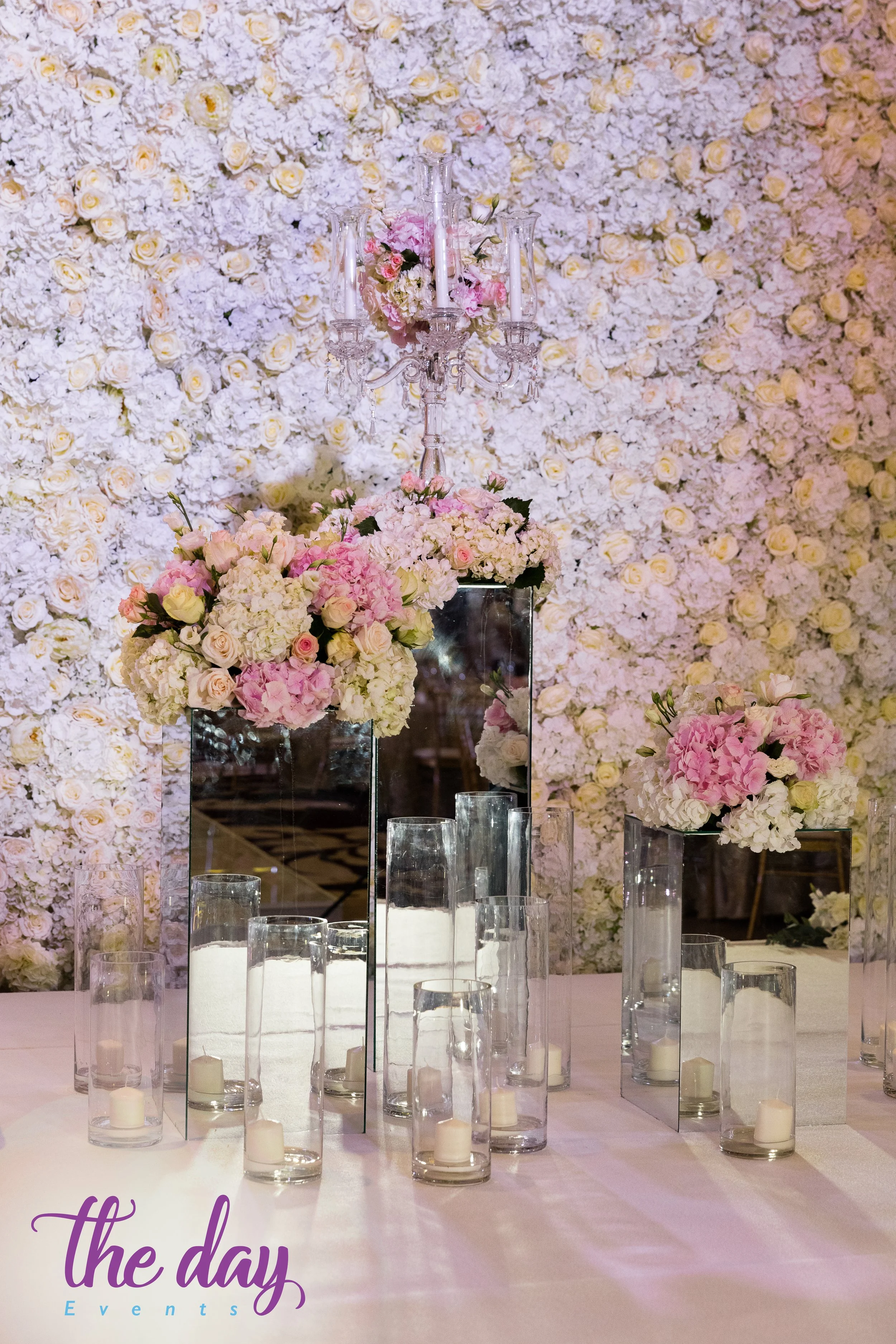 Elegant floral centerpiece with pink and white flowers, surrounded by glass candle holders with white candles, against a wall covered in white roses, at a wedding or event decor setting.
