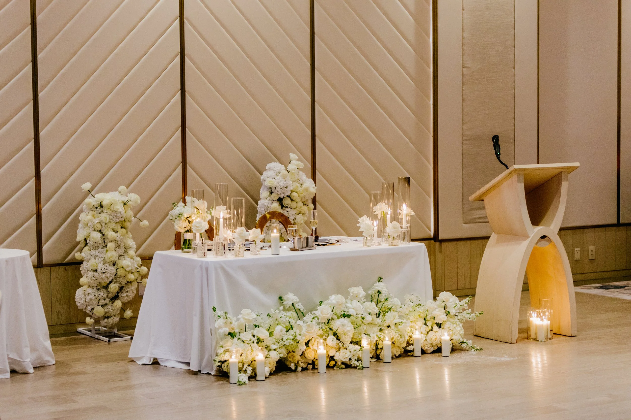 Elegant wedding or event altar decorated with white flowers and candles, featuring a curved wooden podium to the right and a backdrop with striped beige panels.