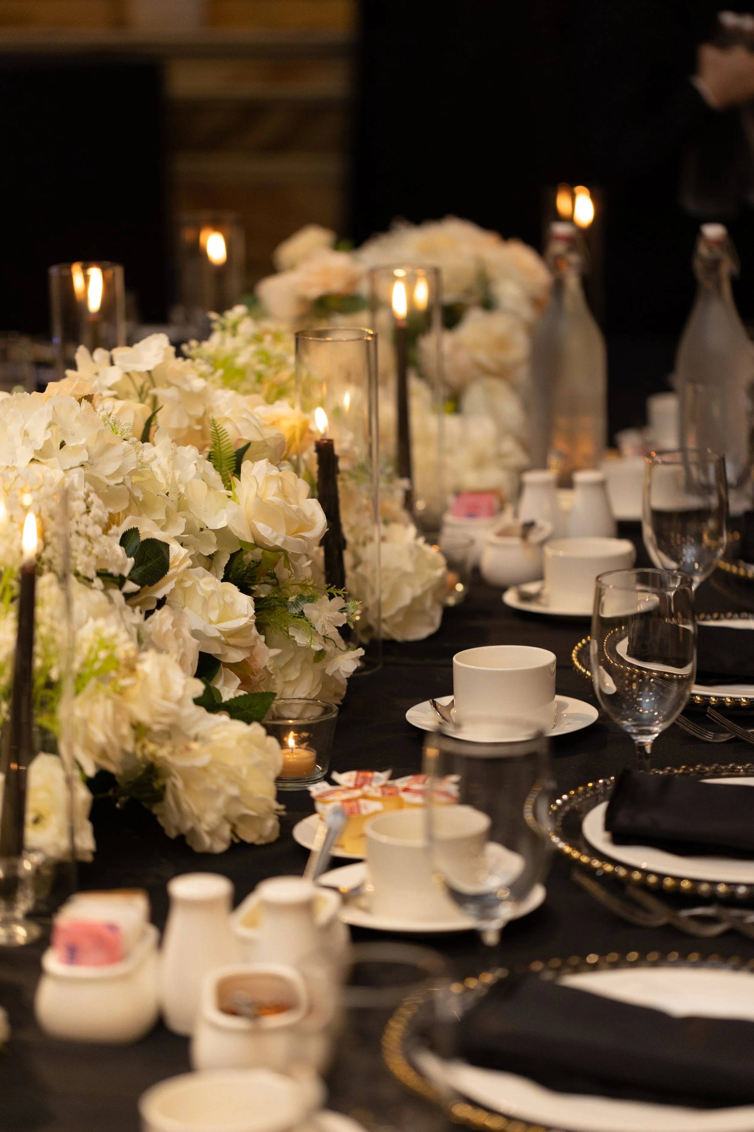 Elegant dining table decorated with white floral arrangements, candles in glass holders, water bottles, glasses, cups, saucers, and black napkins, set for a formal event or celebration.