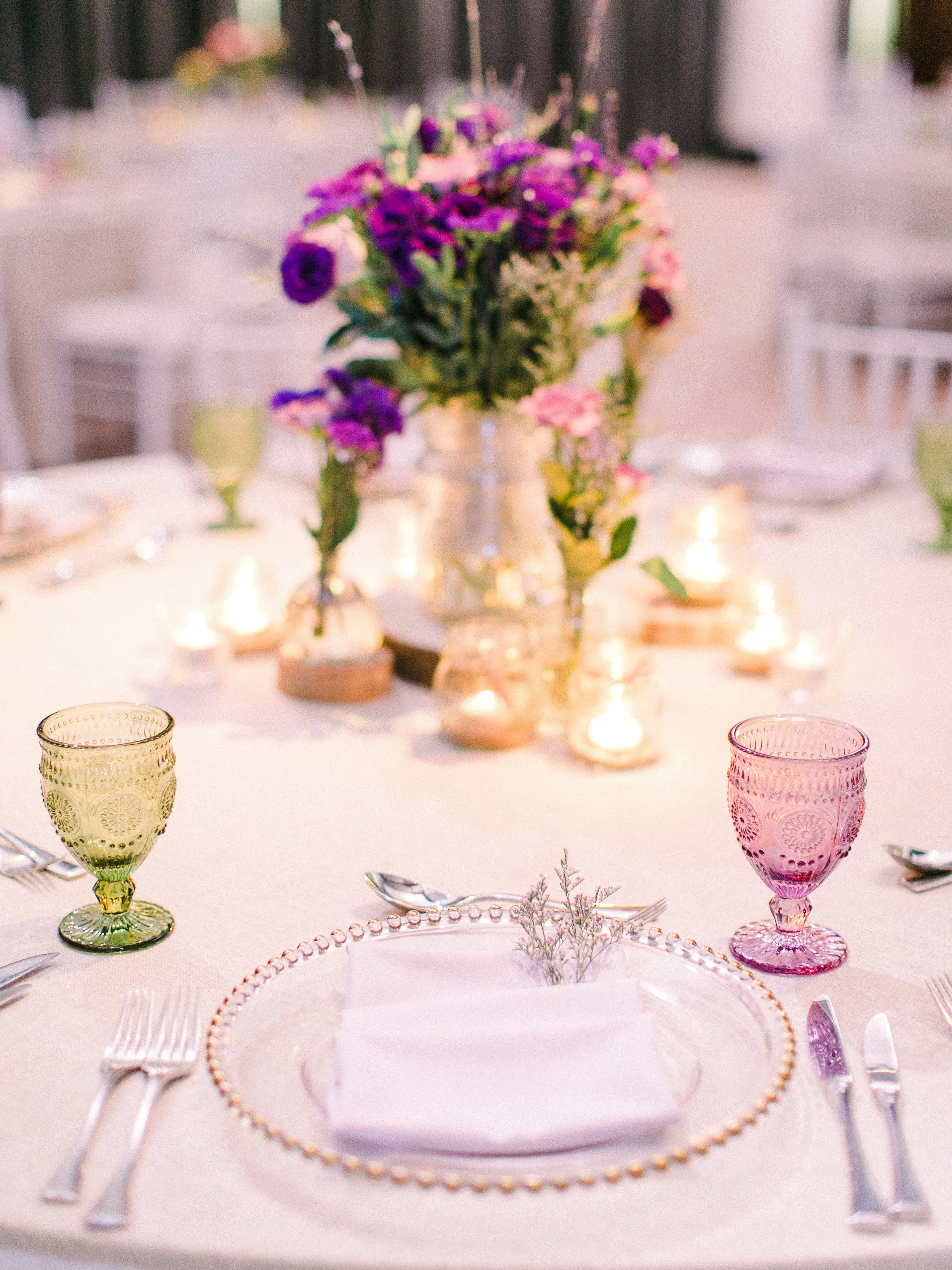 Table setting with colorful vintage glasses, floral centerpiece with purple and pink flowers, and candles, at an outdoor event.