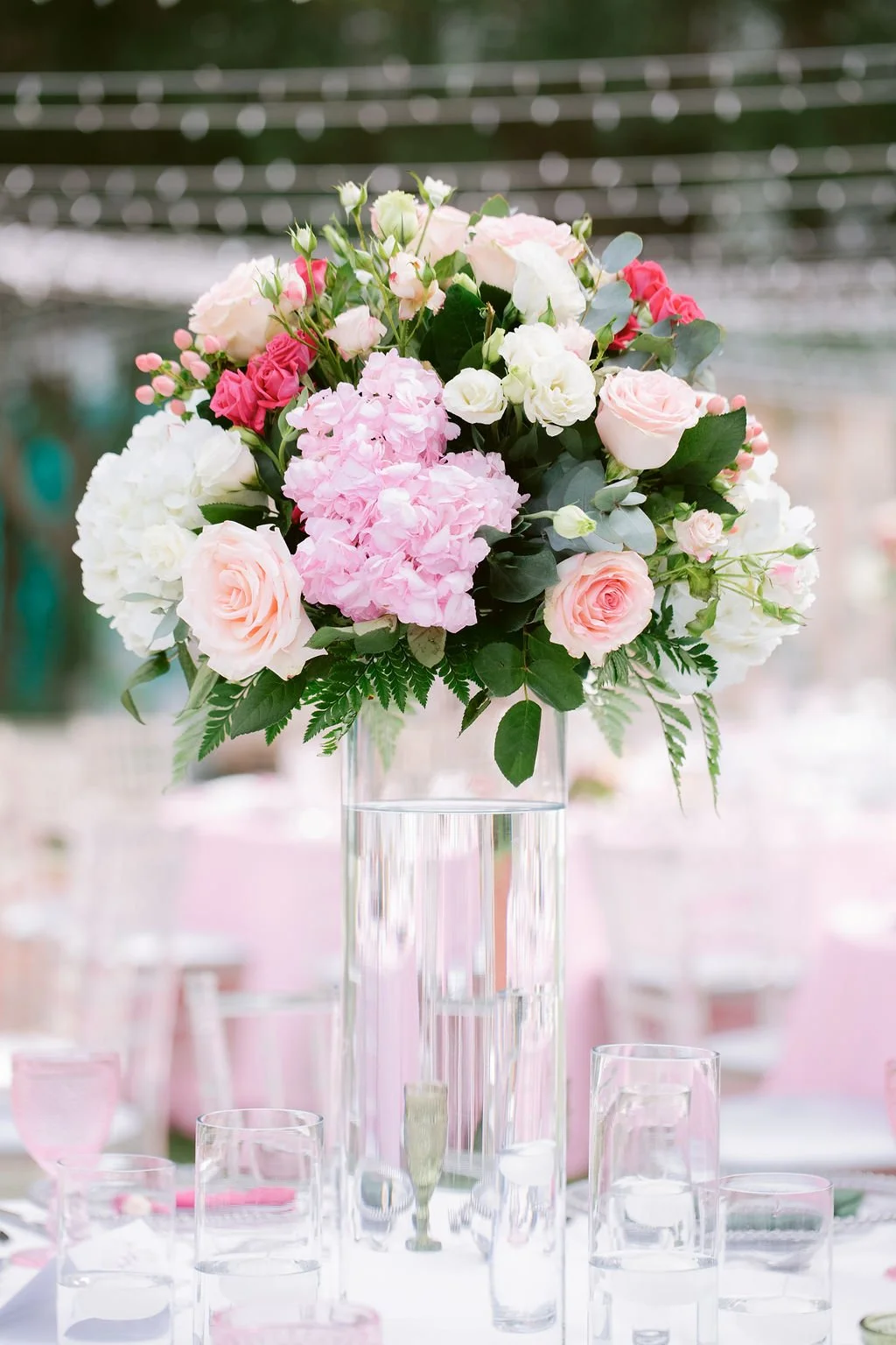 A tall glass vase filled with pink, white, and blush-colored roses, hydrangeas, and greenery serves as a centerpiece at a table with pink and clear glassware.