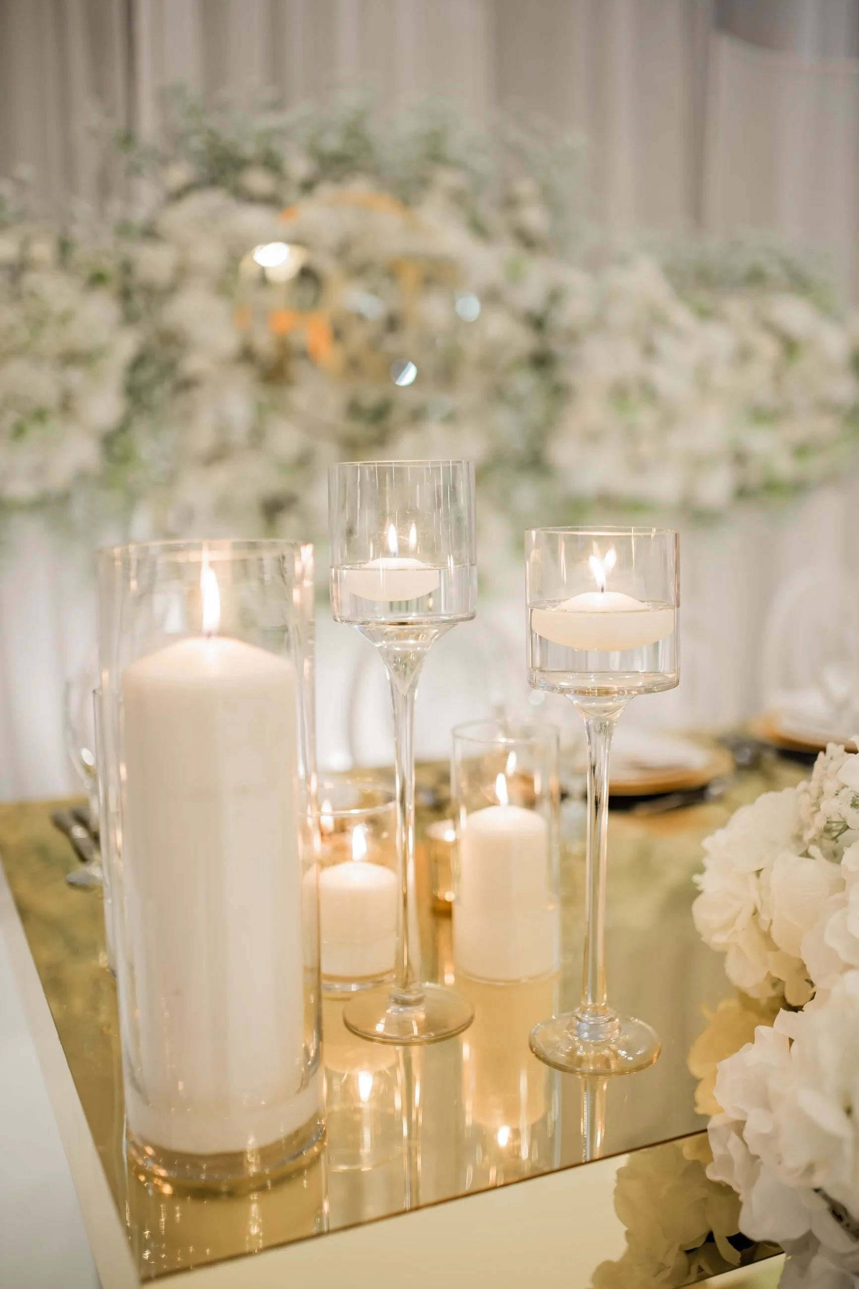 Elegant table centerpiece with tall glass candle holders and white candles, with a blurred floral backdrop.
