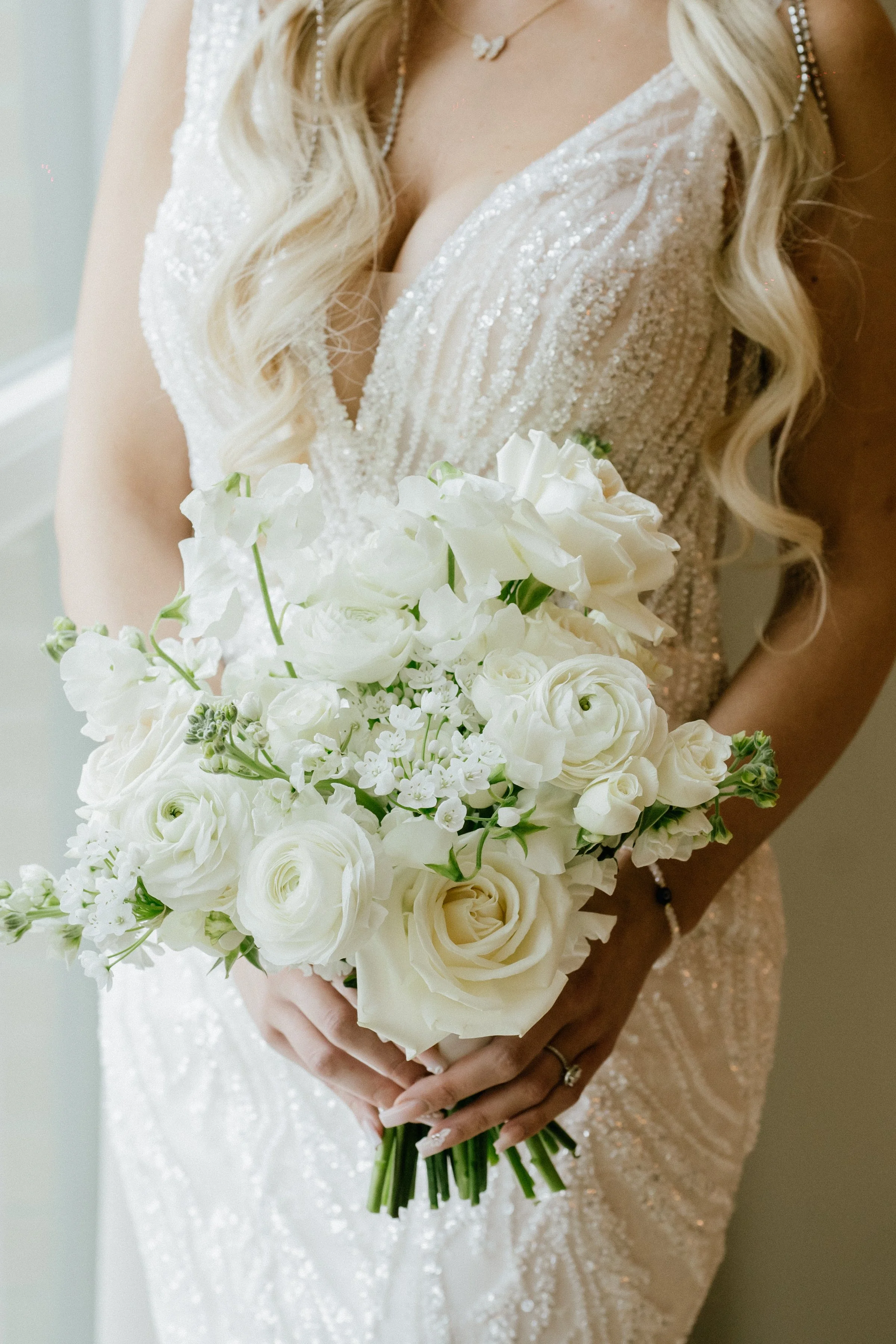 Close-up of a bride holding a white flower bouquet in front of her, wearing a beaded wedding dress and long wavy blonde hair.